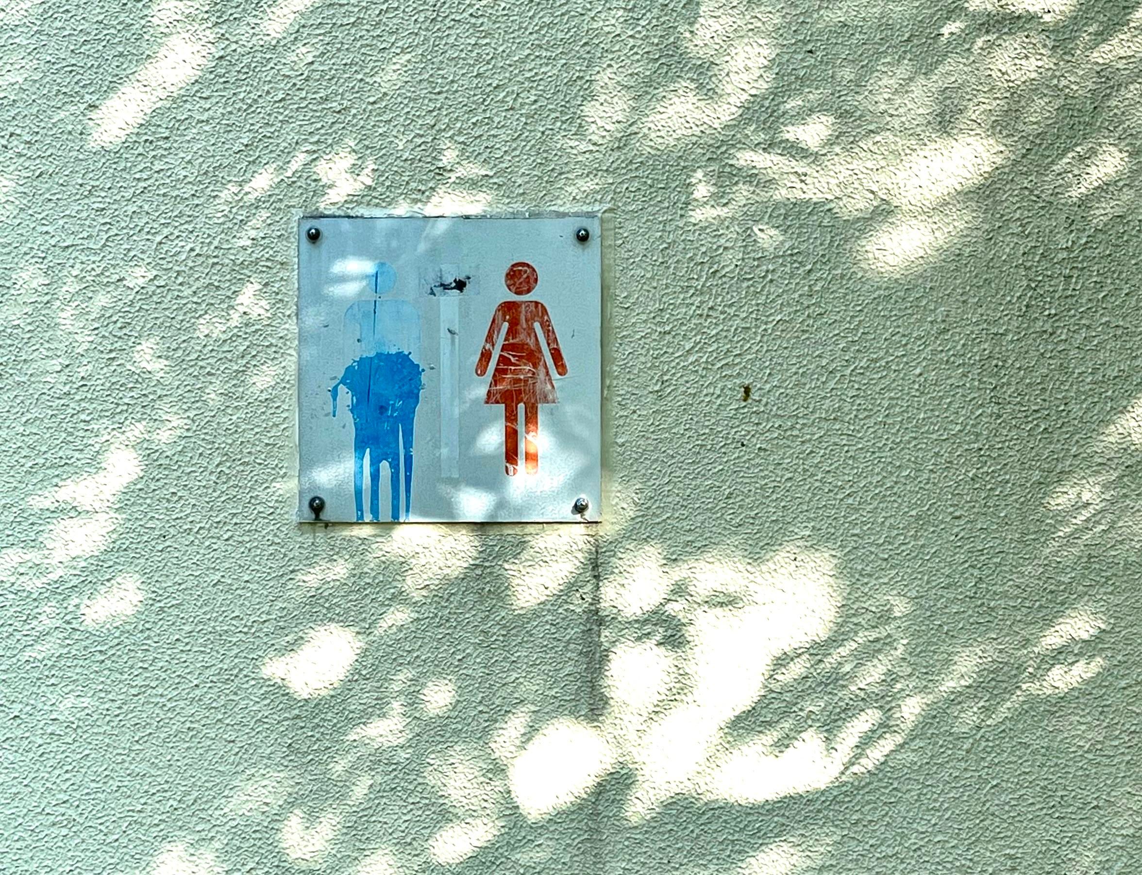 Sur le mur extérieur de toilettes publiques, une petite pancarte avec les pictogrammes pour indiquer le côté "hommes" en bleu et le côté "femmes" en rouge, sauf qu'à la place du petit bonhomme pour les hommes, c'est juste une tache de peinture bleue dégoulinante.