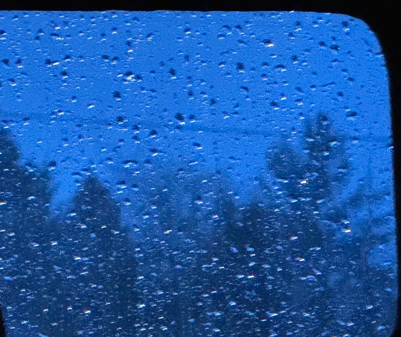 A close-up view of a window with raindrops, showcasing a blue-toned background and blurred silhouettes of trees outside.