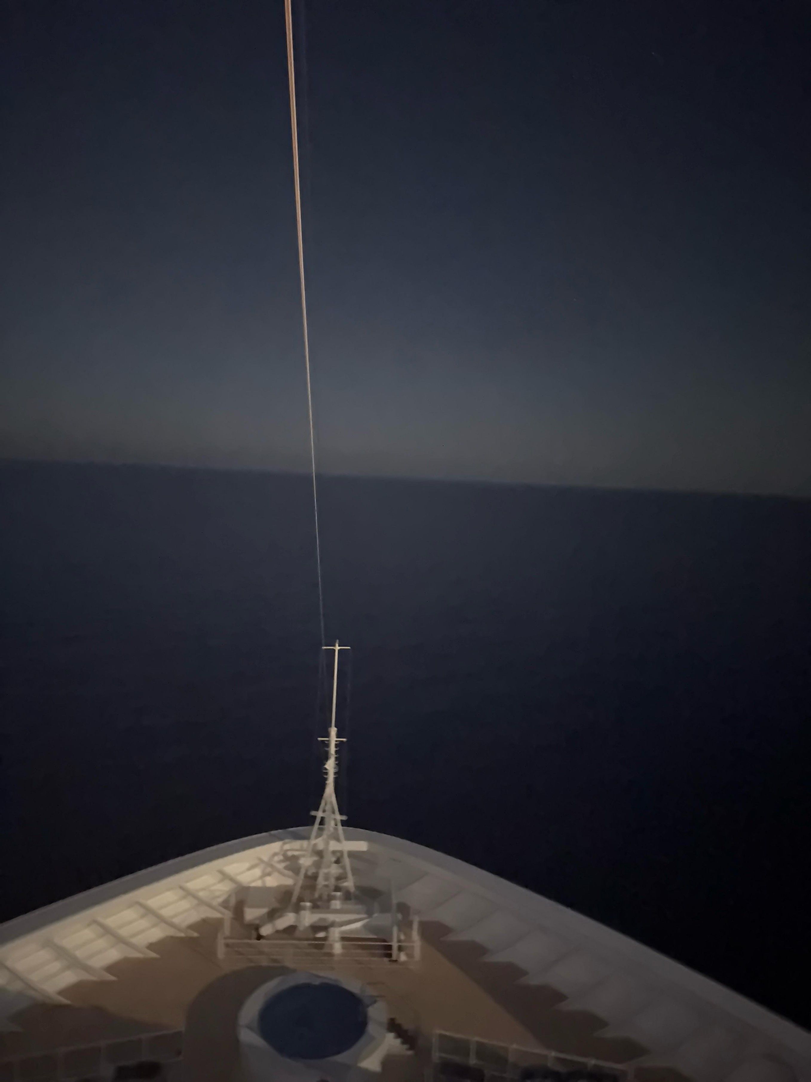 Front deck of a ship at night. The photo is taken from above, showing the pointed bow of the ship heading toward a dark, calm ocean. A tall mast with a line extending upward is in the center. The sky is dark, with a faint light, possibly from the moon or distant horizon, creating a slight gradient above the sea. The scene is quiet and serene, with no visible land or other ships.

Alt-text provided by Be My AI.