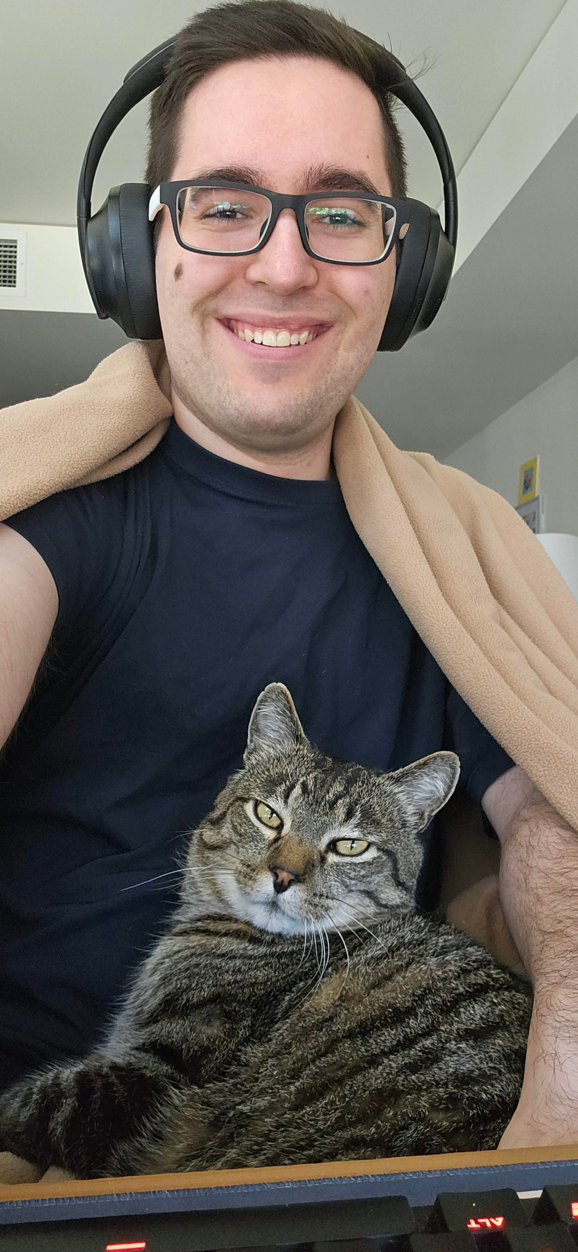 A selfie of me and Momo sitting on my lap. I'm wearing a blue t-shirt and have a sand-colored blanket draped over my shoulders. I have black headphones on. Momo is looking at the camera.
