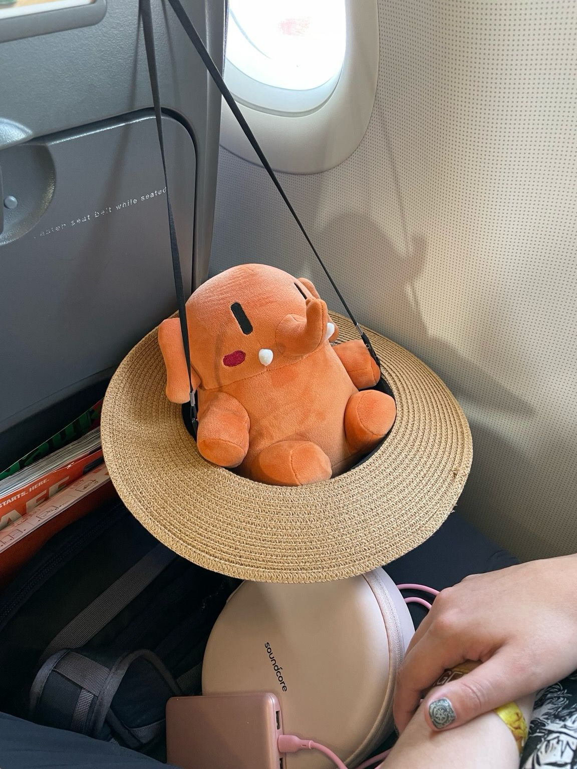 An apricot-colored Mastodon stuffed toy lounging inside a straw hat hanging off an airplane chair. 