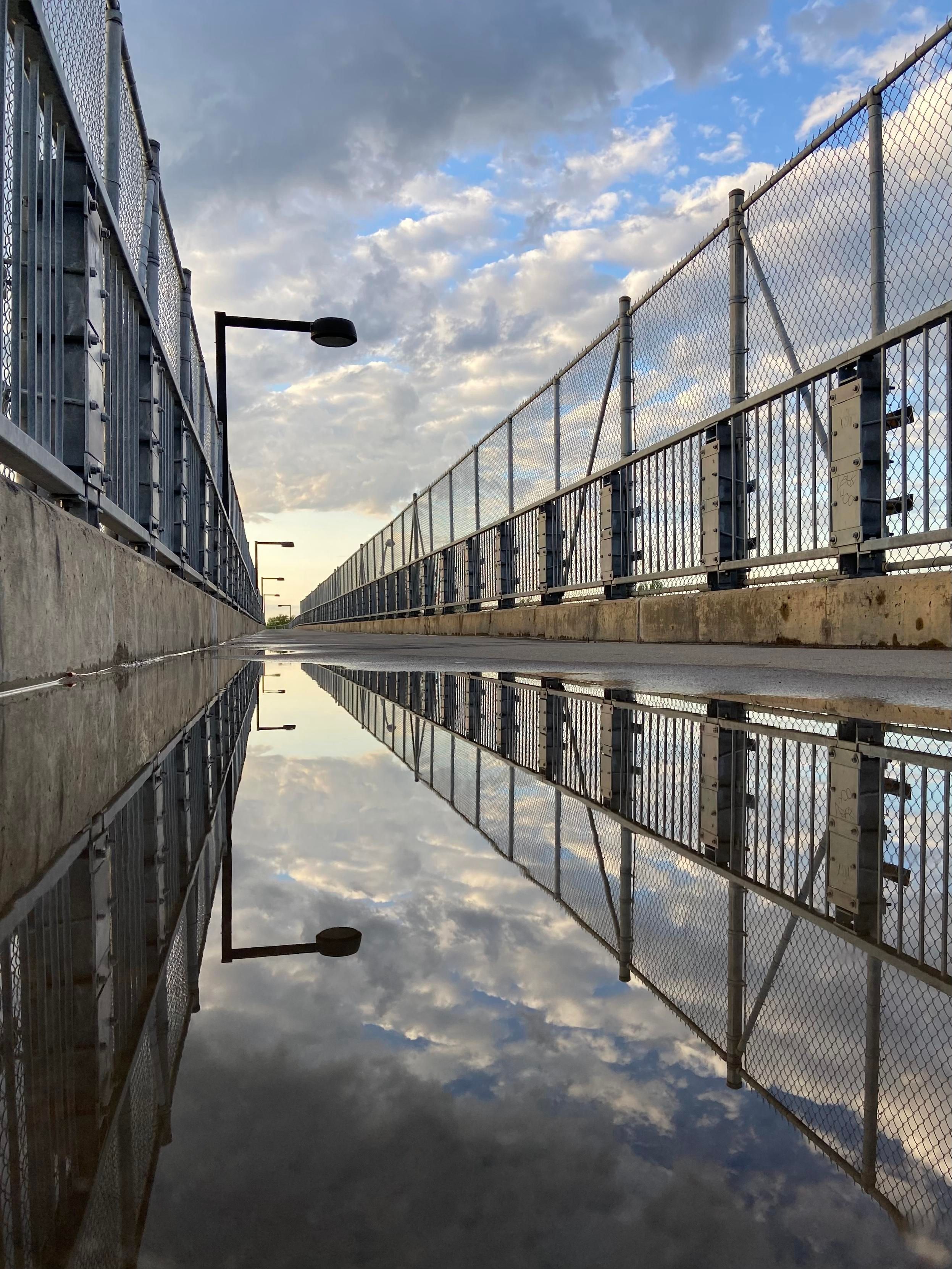 a ground level view of a puddle on a pedestrian bridge, giving a perfect reflection of the concrete sides and metal fences on both sides of the bridge, as well as the partially cloudy sky. 
