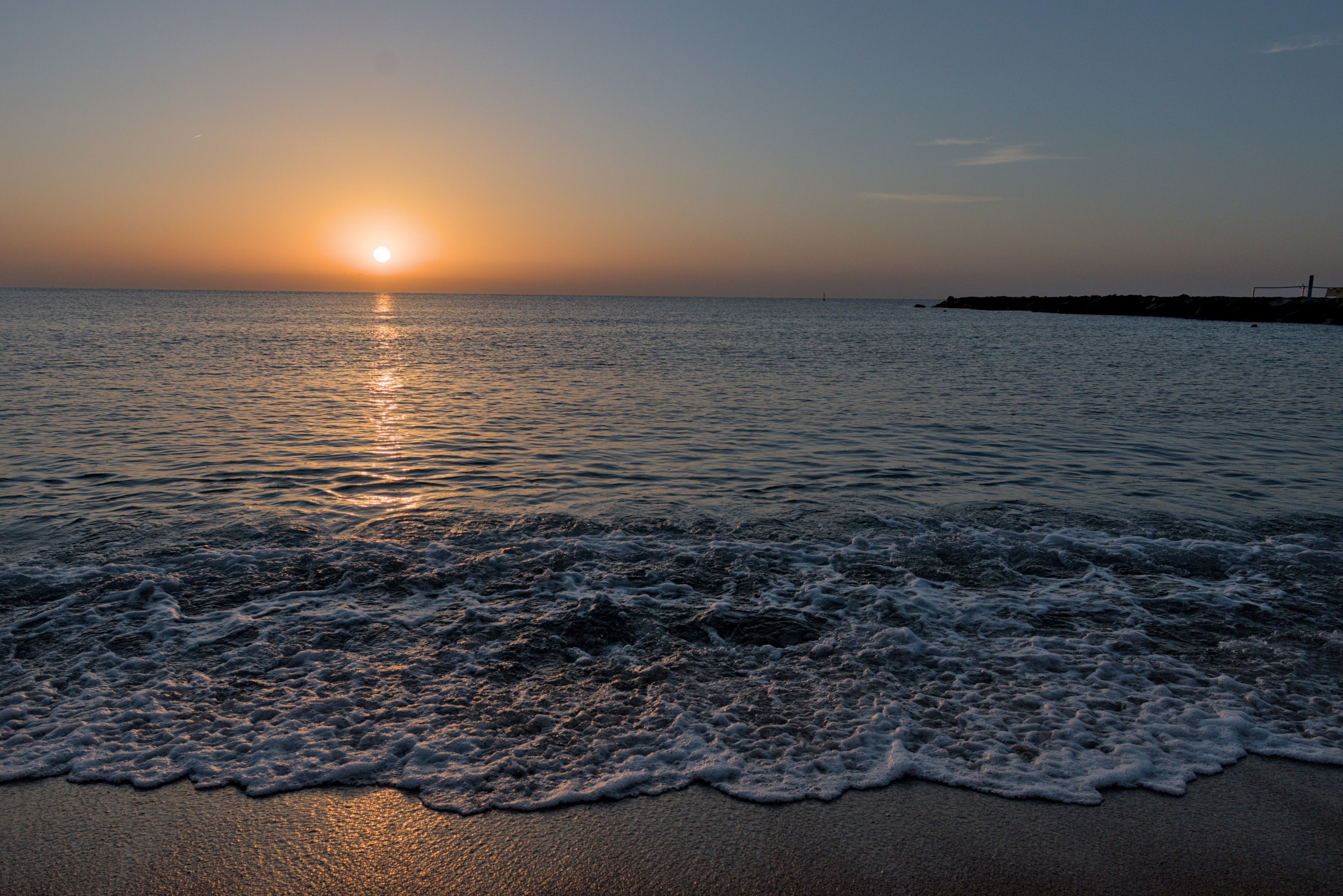 A photo of a sunrise on the beach. The sand itself is visible towards the bottom of the image, quickly replaced by the small waves of a calm see. The sun is only a few degrees above the horizon, and has a long reflection across the water. The sky is a radial gradient around the sun, staring from warm oranges and ending in a dark blue. In the right third of the image, a stone pier can be seen on the line of the horizon.