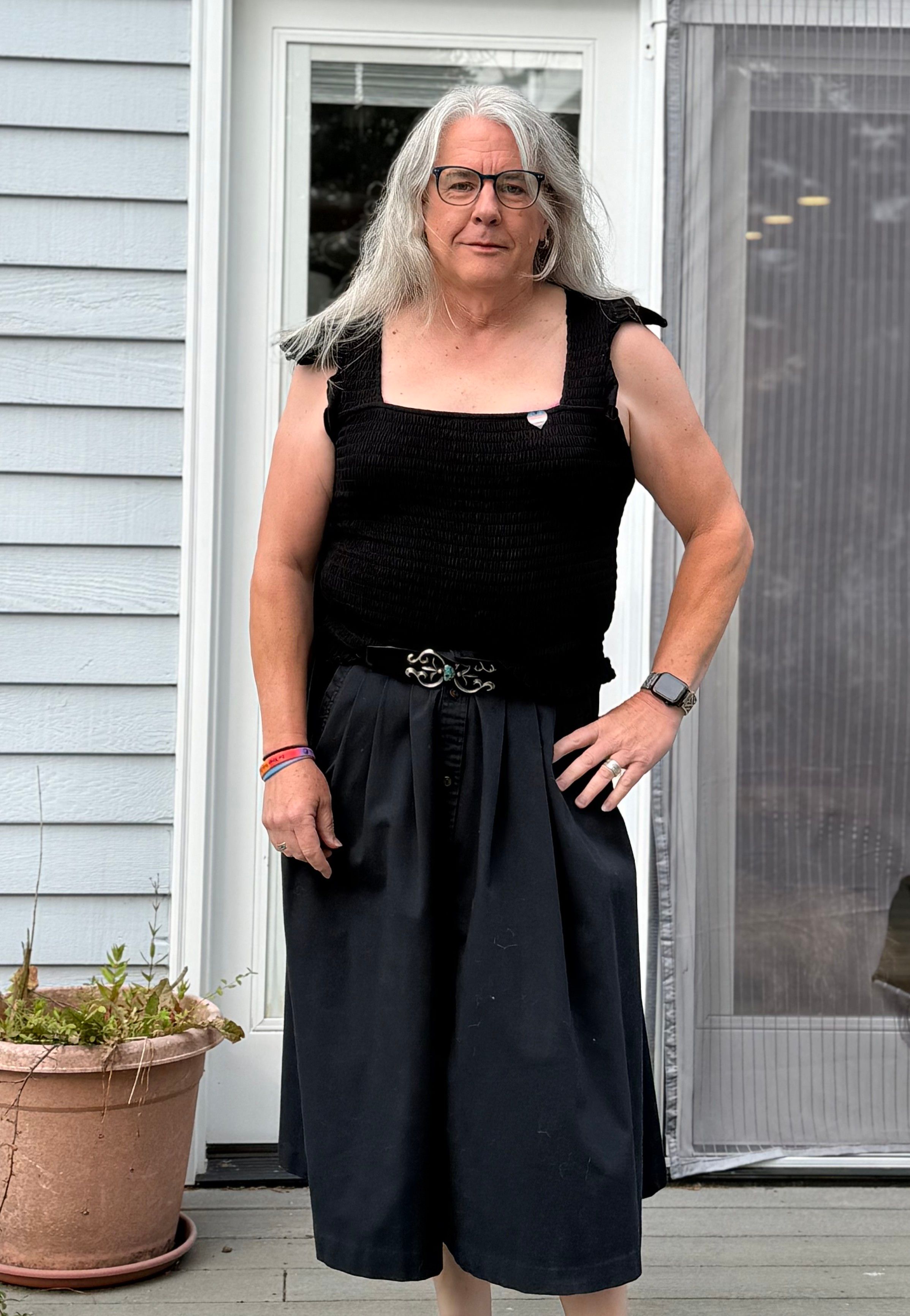 Photo of myself on the deck of a house. Silver hair past my shoulders. Blue/green glasses. A black sleeveless blouse with frilly shoulder straps and a trans heart pin on it. A black ankle-length skirt with a silver and turquoise belt buckle.