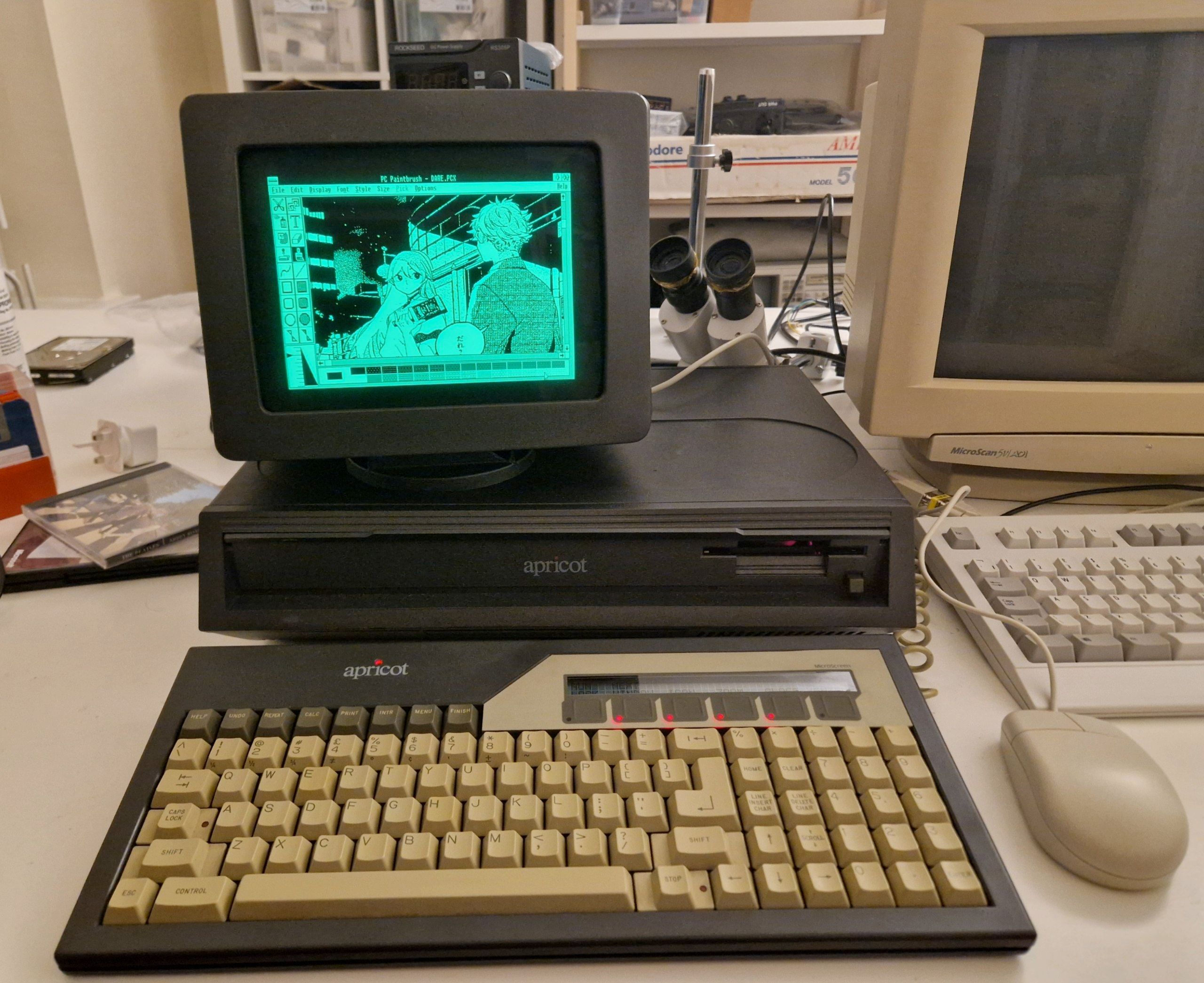A photograph of a vintage Apricot PC computer in dark grey plastic, with a "green phosphor" CRT on top, showing a panel from a comic in Windows Paintbrush. There's a beige mouse to the right of the keyboard, and a larger beige CRT even further to the right.