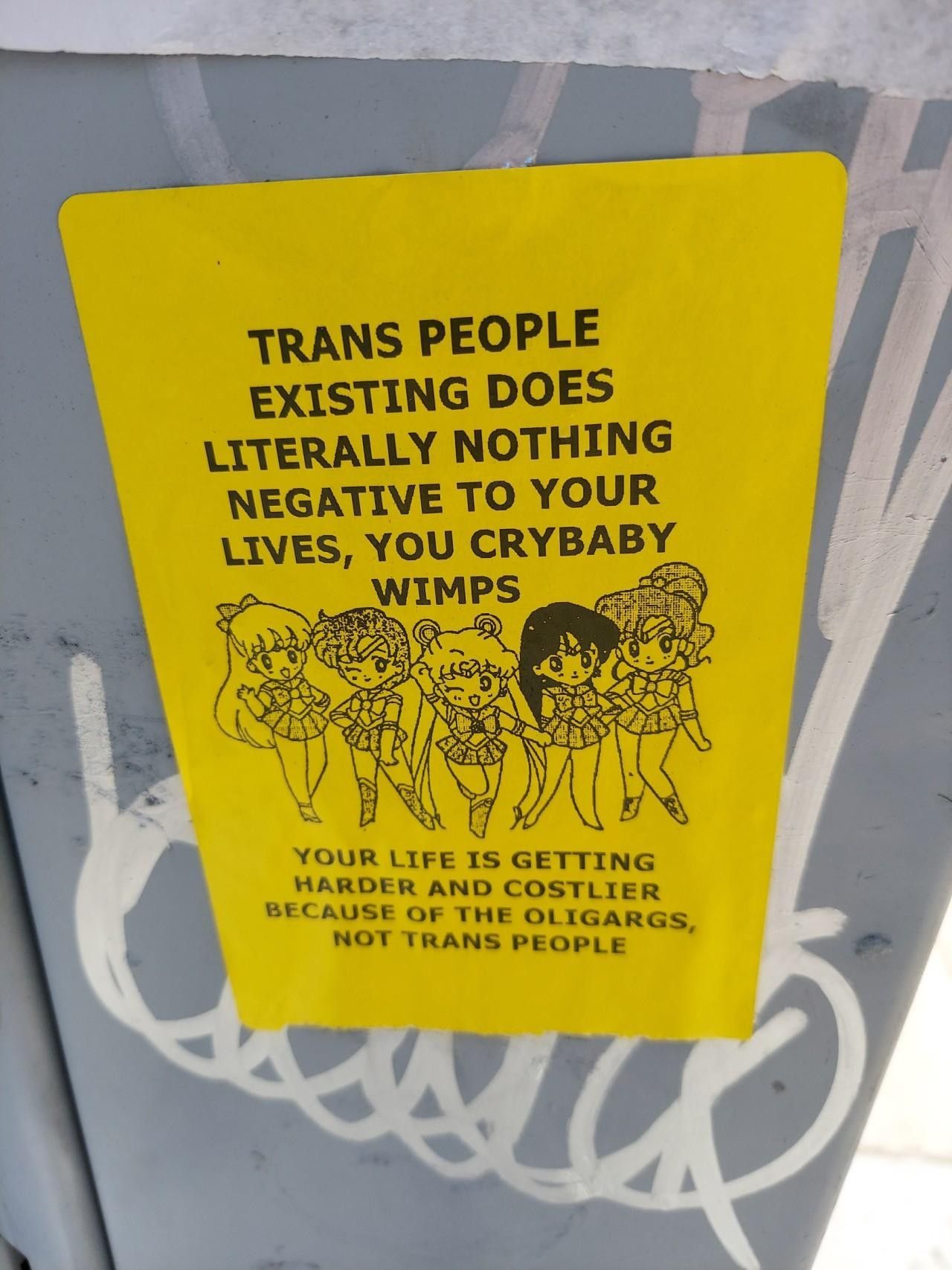 A yellow sticker featuring cartoonish depictions Sailor Moon characters that says, “TRANS PEOPLE EXISTING DOES LITERALLY NOTHING NEGATIVE TO YOUR LIVES, YOU CRYBABY WIMPS. YOUR LIFE IS GETTING HARDER AND COSTLIER BECAUSE OF THE OLIGARCS, NOT TRANS PEOPLE.”