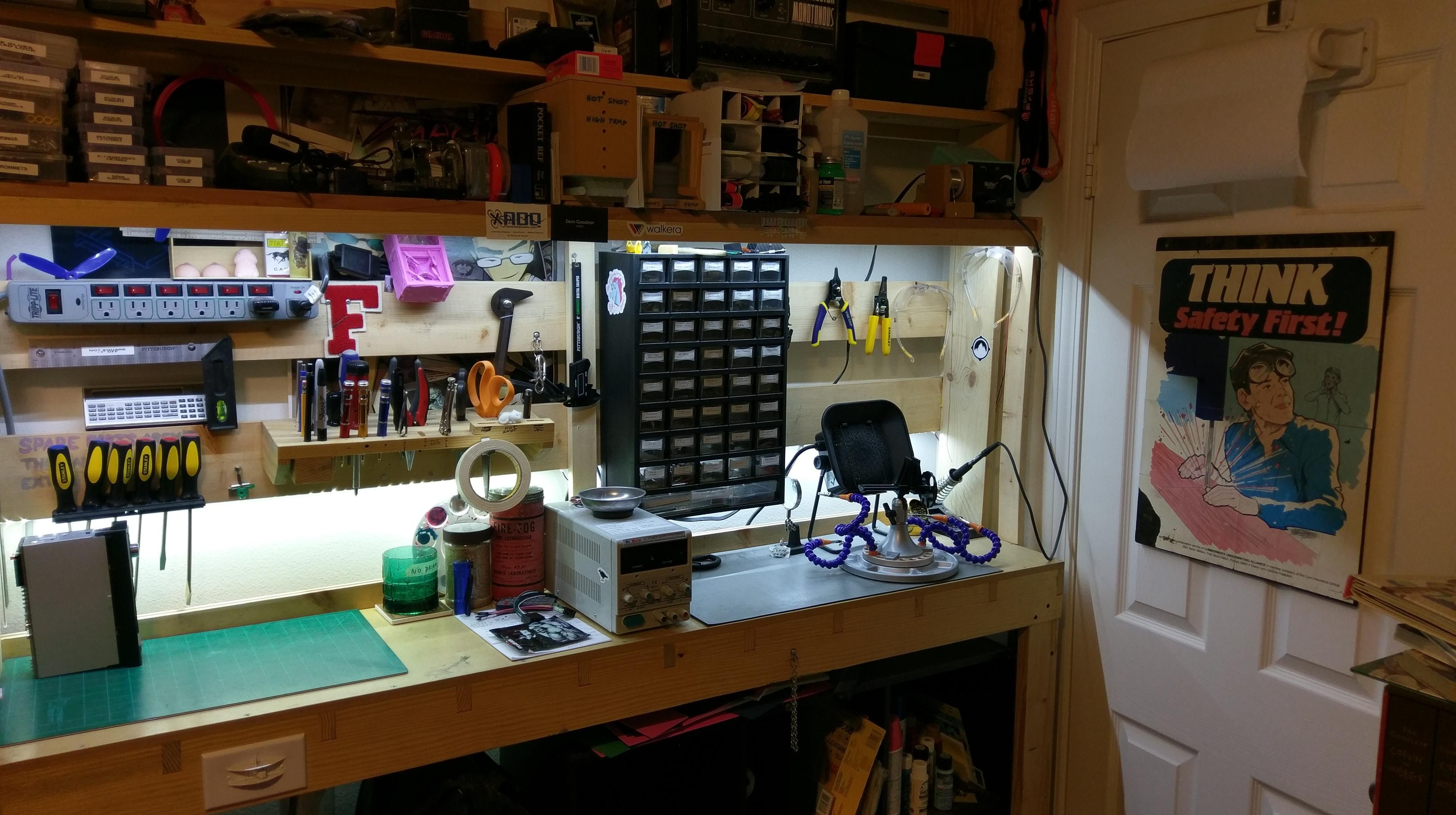 The full uncropped photo of the workbench. It sits in the corner of a bedroom, leaving just enough space for a closet door to open in front of it. The closet door has a paper towel holder and humorous workplace safety sign hanging from it. The sign shows a man with his safety glasses on his forehead, looking off to the side as he uses a band saw. The caption reads "think! safety first!"

The workbench is fairly shallow, and divided into two halves by a small DC power supply in the middle. On the left is a green cutting mat with the tool holder from the other picture mounted above it, as well as various other tools and a surge protector with a red switch over each outlet. Various decorative bits are also resting on the wooden beams. The right side has a gray ESD mat, with a Panavise, fume extractor, and soldering iron pencil on it, with a cord leading up to the iron's base on the shelf above, next to a spool of solder. The rest of the overhead shelves are packed end to end with various tools, boxes of screws, hot glue, heat-shrink tube, and other similar items. The entire bench is lit by LED lights set in the underside of the shelf, activated by a switch on the front edge of the benchtop. More shelves are barely visible under the bench, which hold even more tools, supplies, and various maker ephemera.