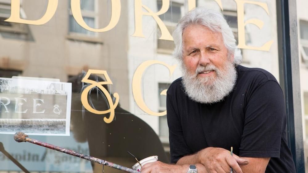 Portrait of a man posing in front of a sign he is painting on a window. He is holding his sign painting materials in his hands.