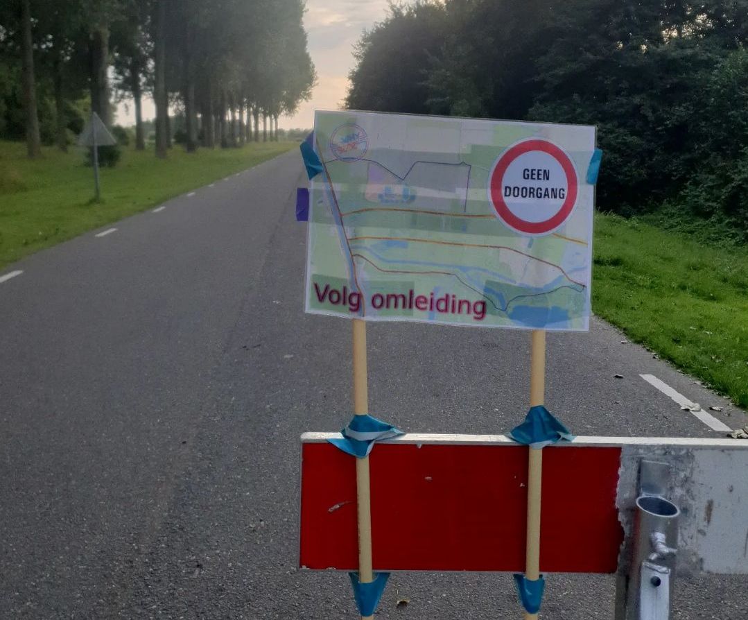 A road with a barrier blocking entry to the right lane, and a sign taped to it with the WHY logo, A "geen doorgang" symbol, the words "Volg omleiding", and a map showing a rather long diversion route from the path to the eastern entrance (where this sign is), around the event location, to the car entrance in the north.