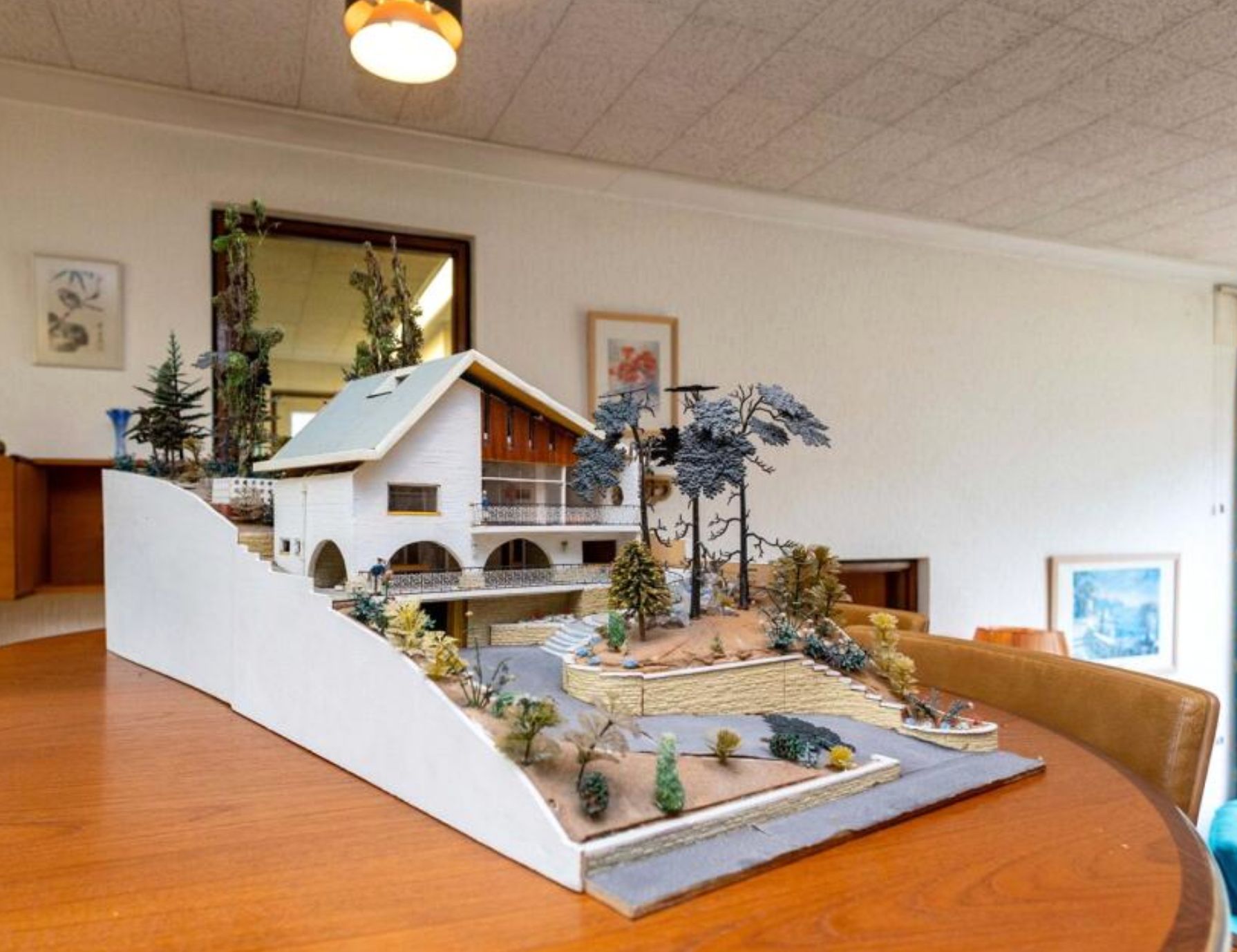 A model of the house sitting on the dining room table. It's on a sloped block of land with a winding driveway, and the house is 3 stories and in a style from the 1970s. It's a very detailed model with trees and gravel. 