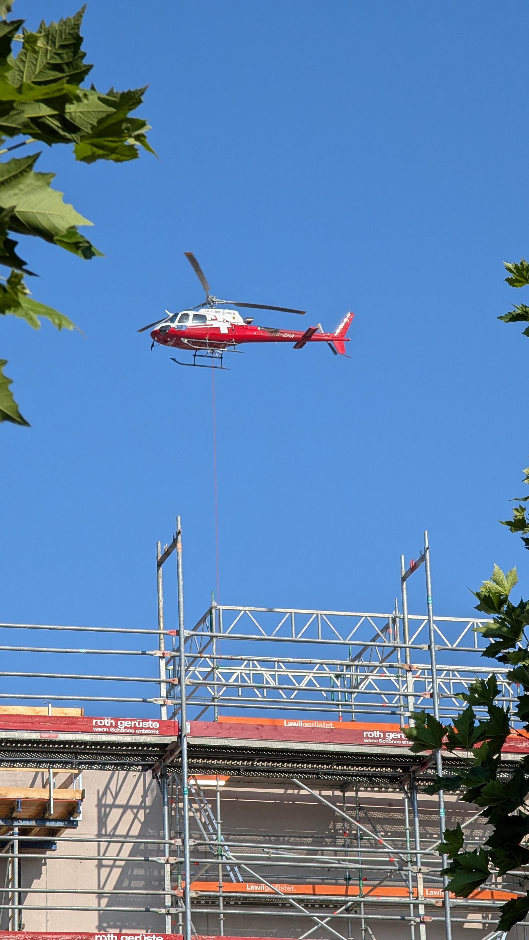 Ein rot-weisser Helikopter schwebt über einer Baustelle. An ihm hängt unten ein Seil, mit dem er wohl Dinge anliefert. Was daran häng, sieht man nicht, es ist vom im Bau befindlichen Gebäude und dem Gerüst daran verdeckt. Der Himmel ist blau, am Bildrand sieht man ein paar Blätter einer Platane.