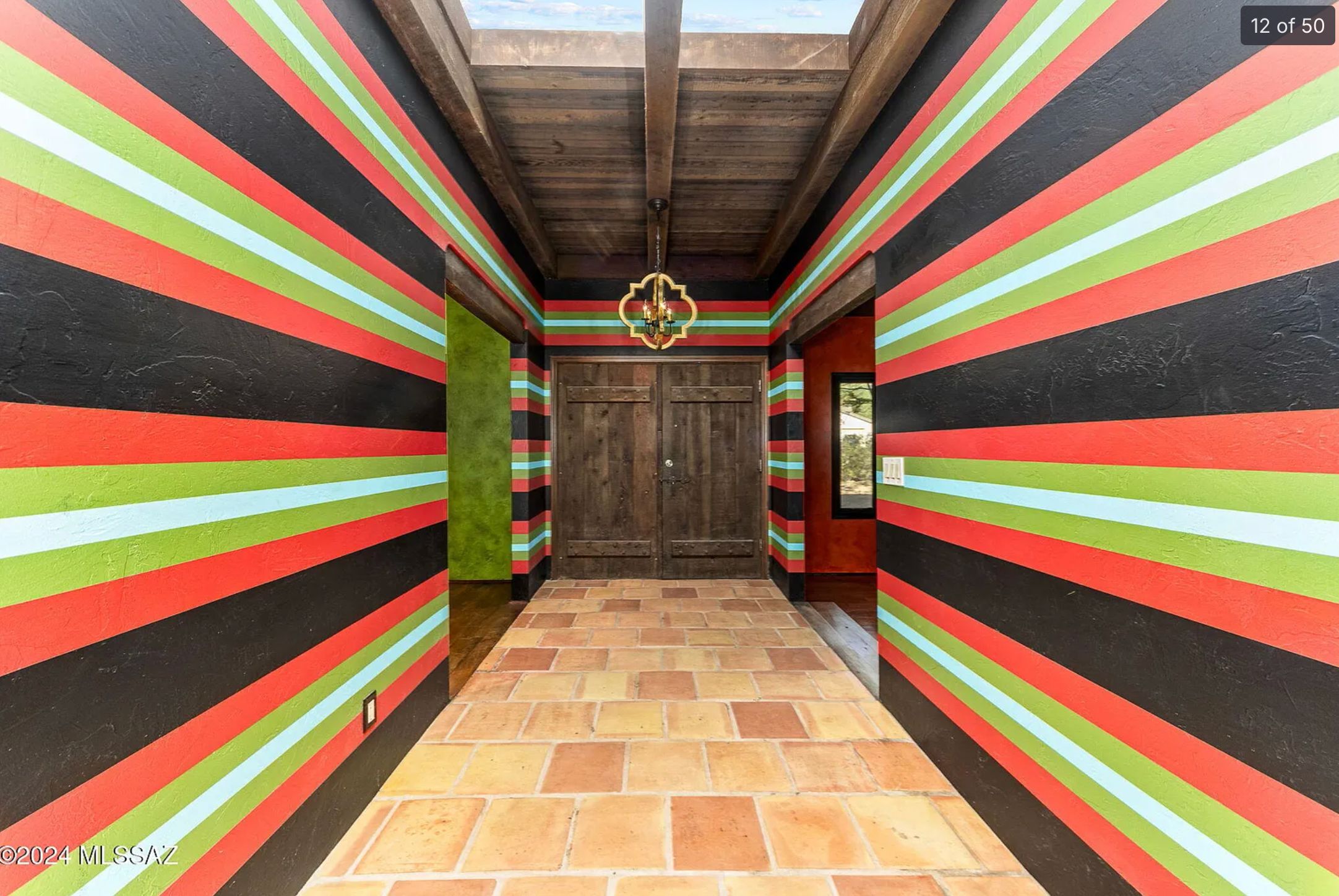 A hallway leading to a front door. The wallpaper is very bright with horizontal stripes of orange, lime green, white and black. The floor is pale orange tile and the door and ceiling are dark coloured wood. 