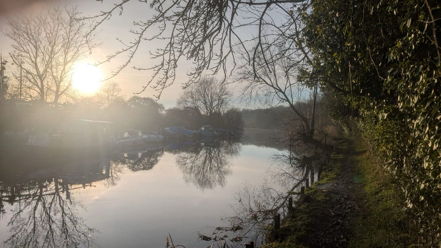 Grove Ferry near Canterbury UK. Sun is just about to set over the river, and the water is as still and reflective as glass. There are river boats moored to the left of the river, and a narrow muddy path winds off into the distance following the edge of the river. The path is walled to the right with trees and hedgerows. Talking of leaning to the right, if Kier Starmer was walking towards you on this narrow riverside path, with no one else in sight, you could definitely push him in. Just saying.