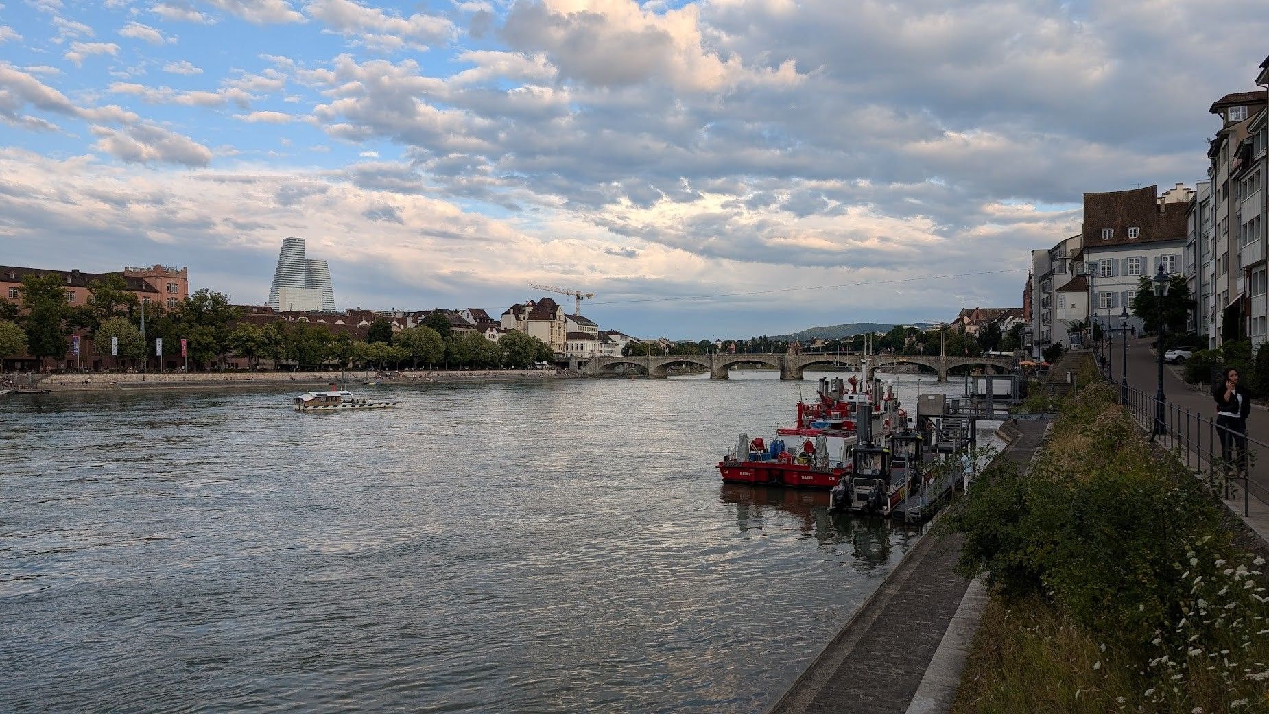 Photo of the view from the promenade above the river Rhine over the river and the northern part of Basel ("Kleinbasel") with two big white chunky towers. The sky is somewhat cloudy. There is a firefighter boat in the foreground on the right.