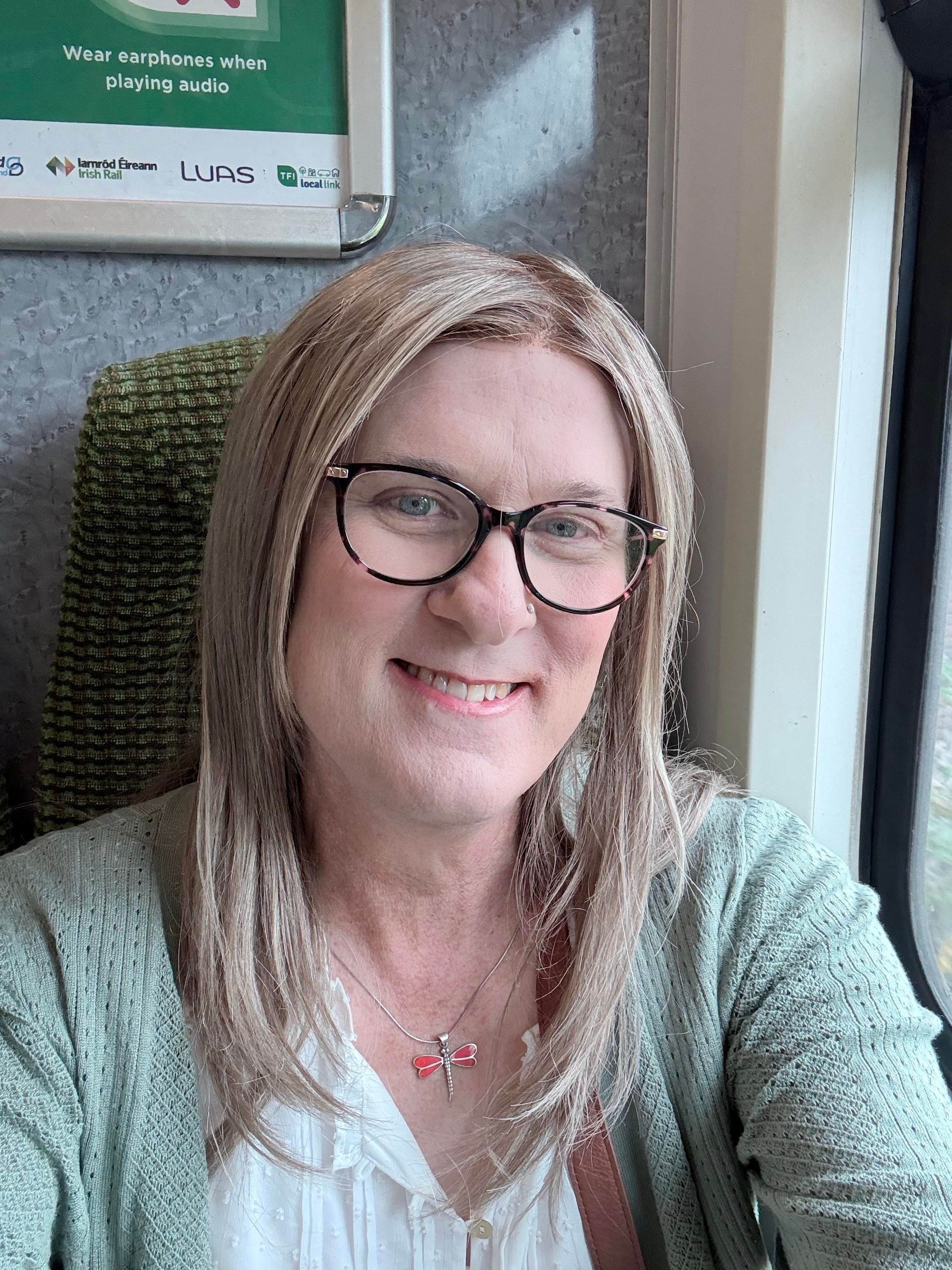 Sophie has longer than shoulder length blonde hair, is wearing a green cardigan over a white top, and a red dragonfly necklace. She has pink and black tortoiseshell glasses. She is taking the DART train in Dublin for the first time!