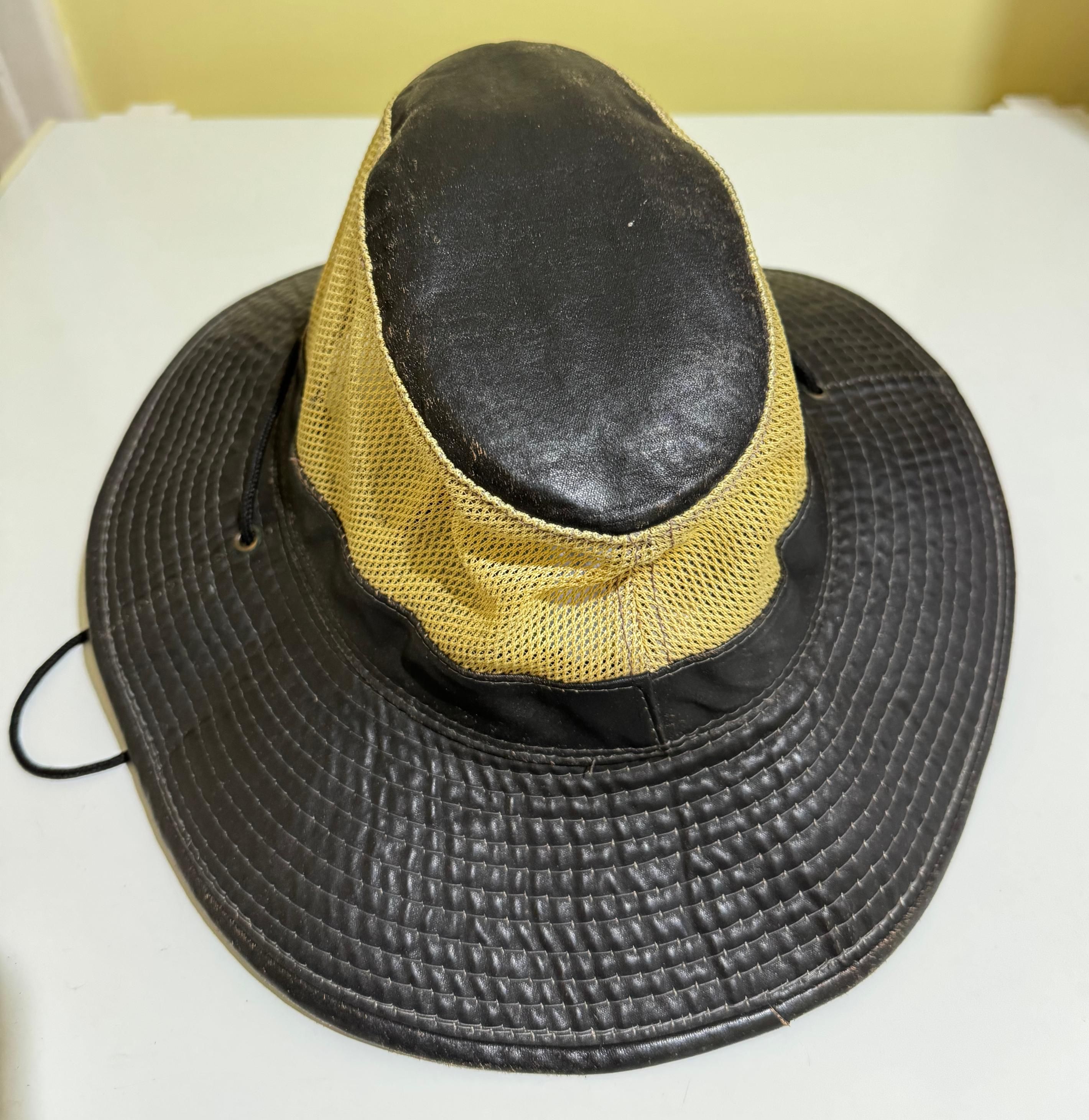 A dark brown floppy hat with gold mesh sides sits on a white surface. The hat looks woven due to the stitching keeping the layers together. There's a long black bootlace threaded through two holes in the brim, forming the neck strap.