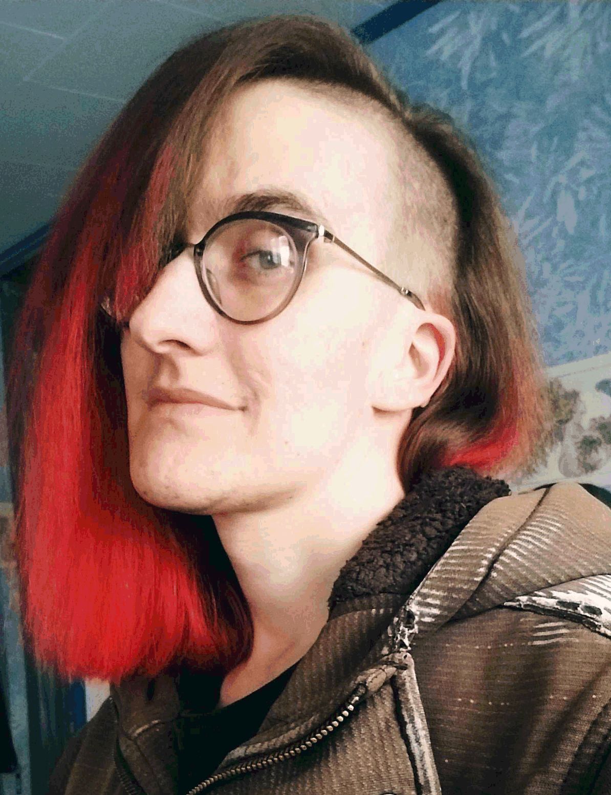 A genderqueer enby transfem with brown and red hair looks at the viewer with a confident smirk. She's wearing round glasses with blue frames and a UV-burned black hoodie. Pronouns are it/its, they/them, she/her.