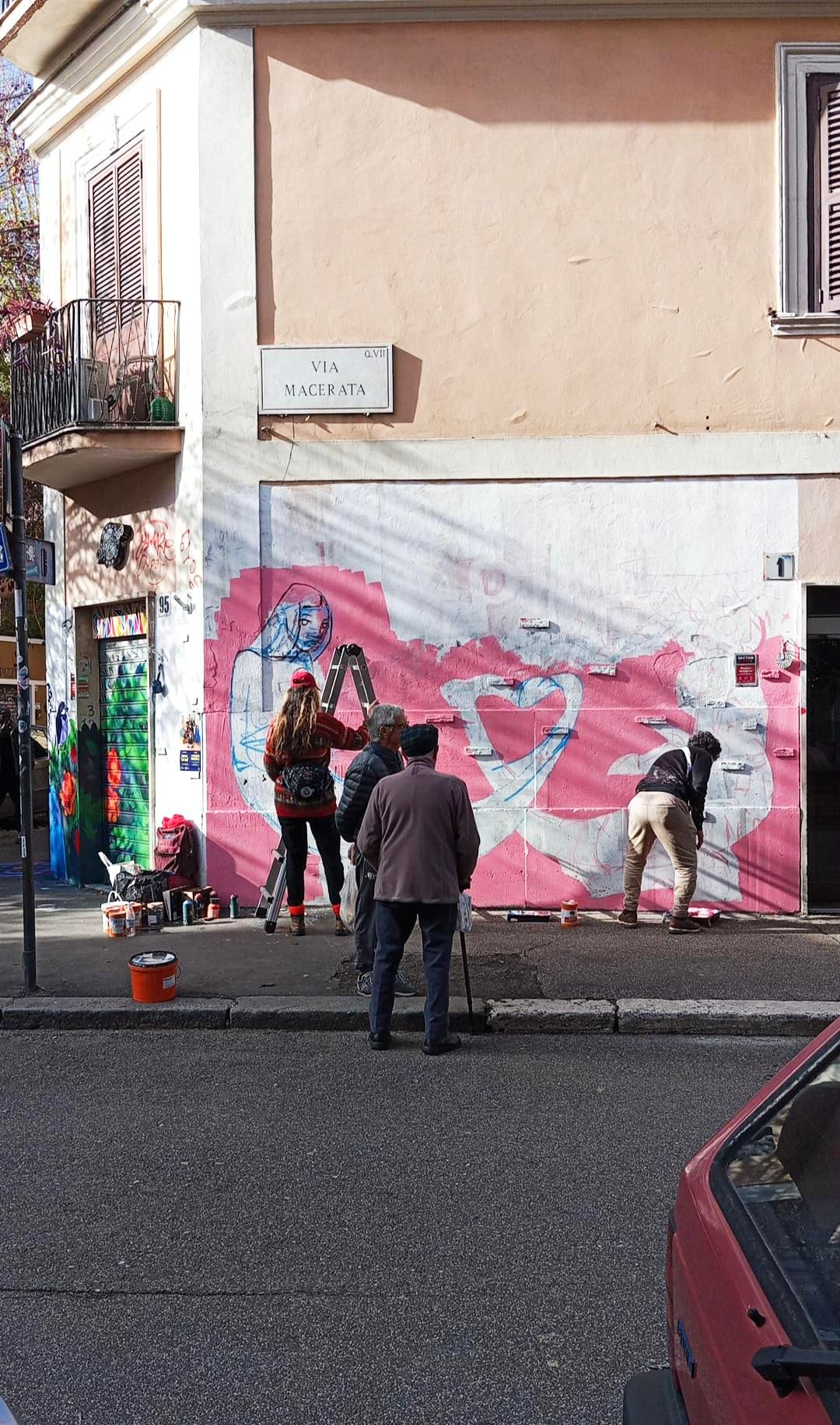 Delle ragazze dipingono un murale, a guardarle due anziani col cappello. In primo piano il retro di una Panda rosso scuro