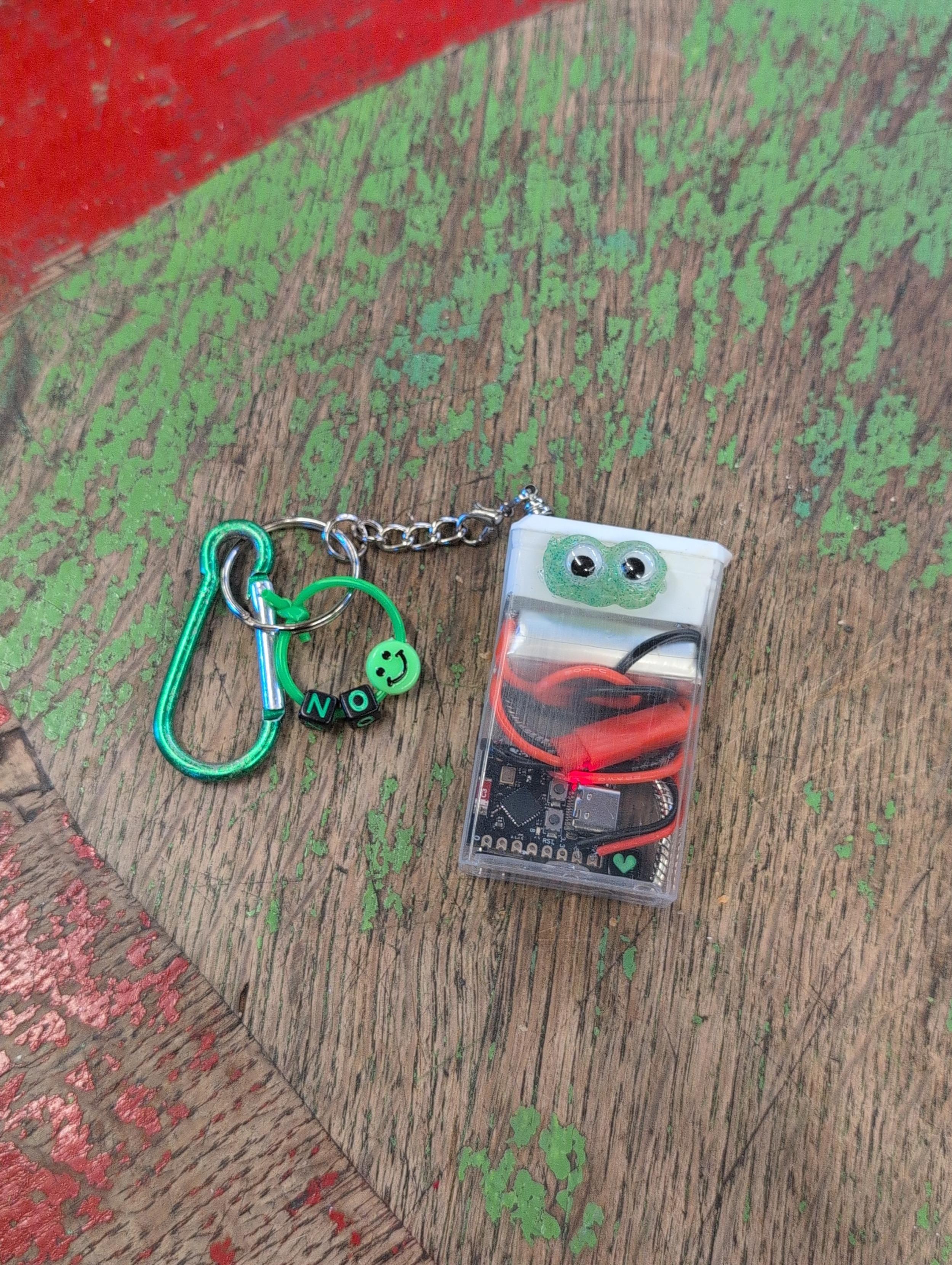 server charm: a microcontroller and a battery inside a tictac box with googly eyes. attached is a keychain with green dangly items.