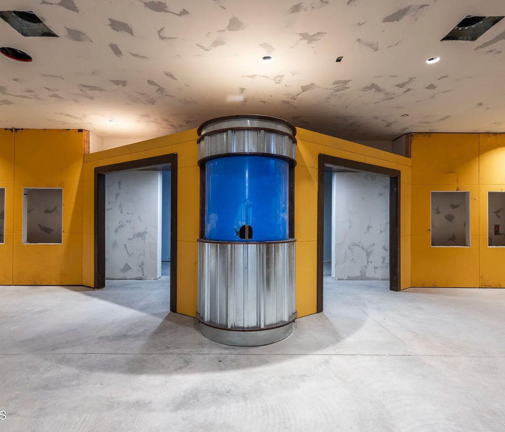 A yellow room with angled doors and in the middle is a silver and blue circular structure that might be a fish tank. The ceiling is unfinished and the walls are bright yellow. 