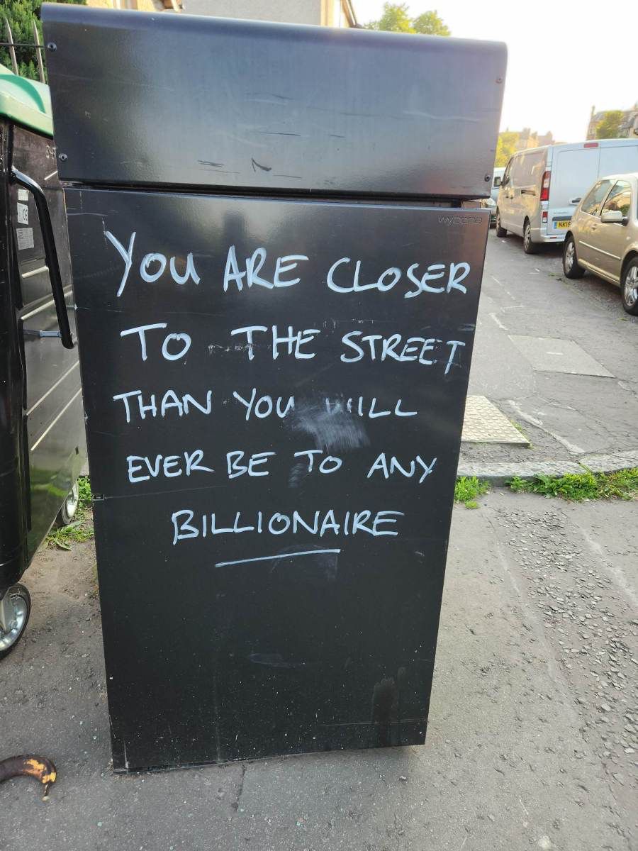 Bild einer schwarzen Säule an einer Straße. Auf der Säule steht in weißen Großbuchstaben : „you are closer to the street than you will ever be to Any billionaire“