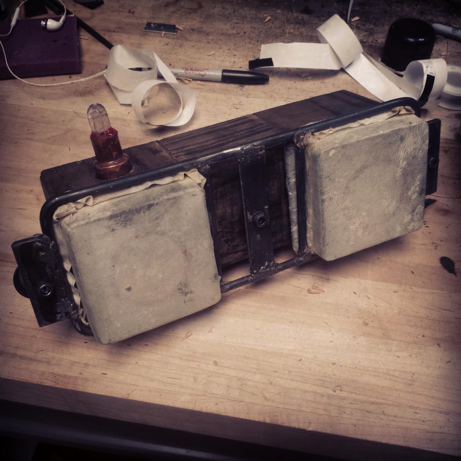 Closeup of the small boombox. A wooden box with a steel frame that tensions two square drumskins on the front. A quartz point in a copper socket acts as an indicator lamp.