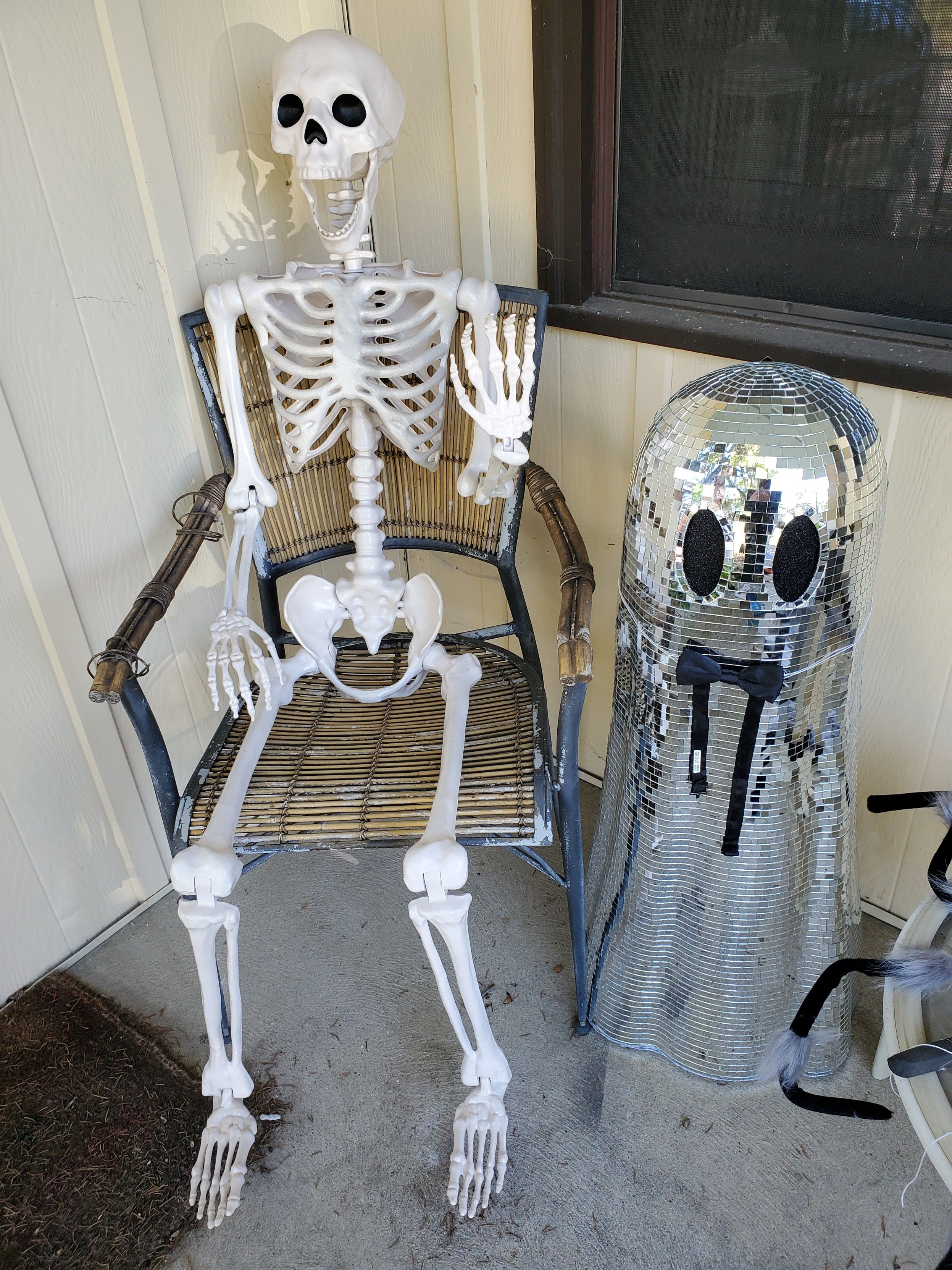 A nude plastic skeleton in a chair, and a mirrored ghost statue with a black bow tie, both sitting on my porch.