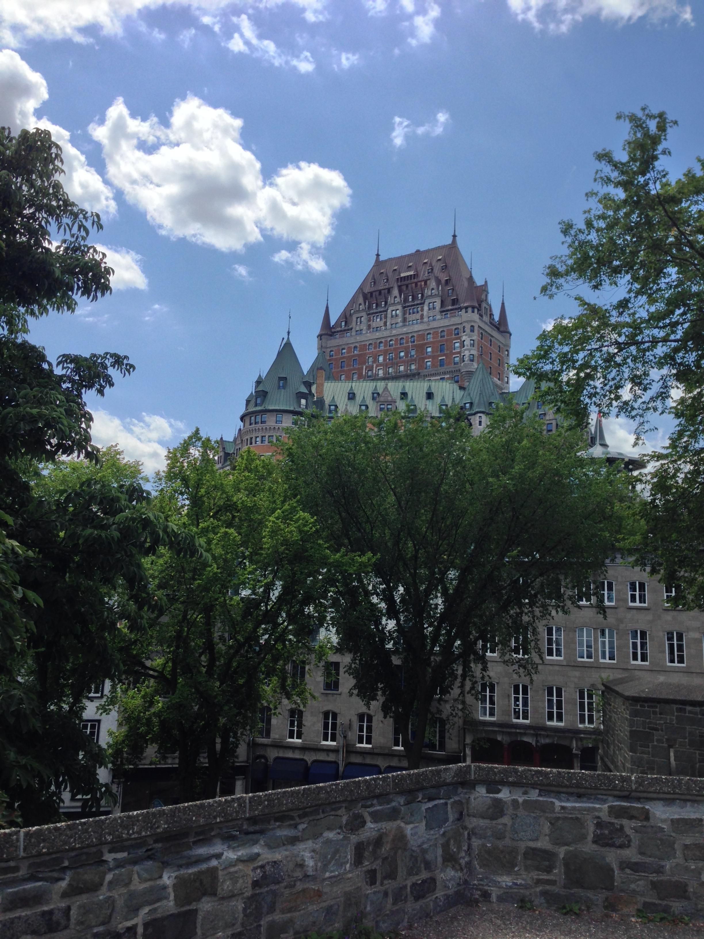 The Château Frontenac Hotel on top of a hill. The roof is copper brown, with small spires peeking out. There are trees below and on the sides covered in leaves, hiding the buildings behind them.

There’s a stone wall at the front and blue cloud-covered sky.
- - -
L'hôtel Château Frontenac au sommet d'une colline. Le toit est brun cuivre, avec de petites pointes qui sortent. Il y a des arbres en dessous et sur les côtés couverts de feuilles, cachant les bâtiments derrière eux.

Il y a un mur de pierre à l'avant et un ciel bleu couvert de nuages.