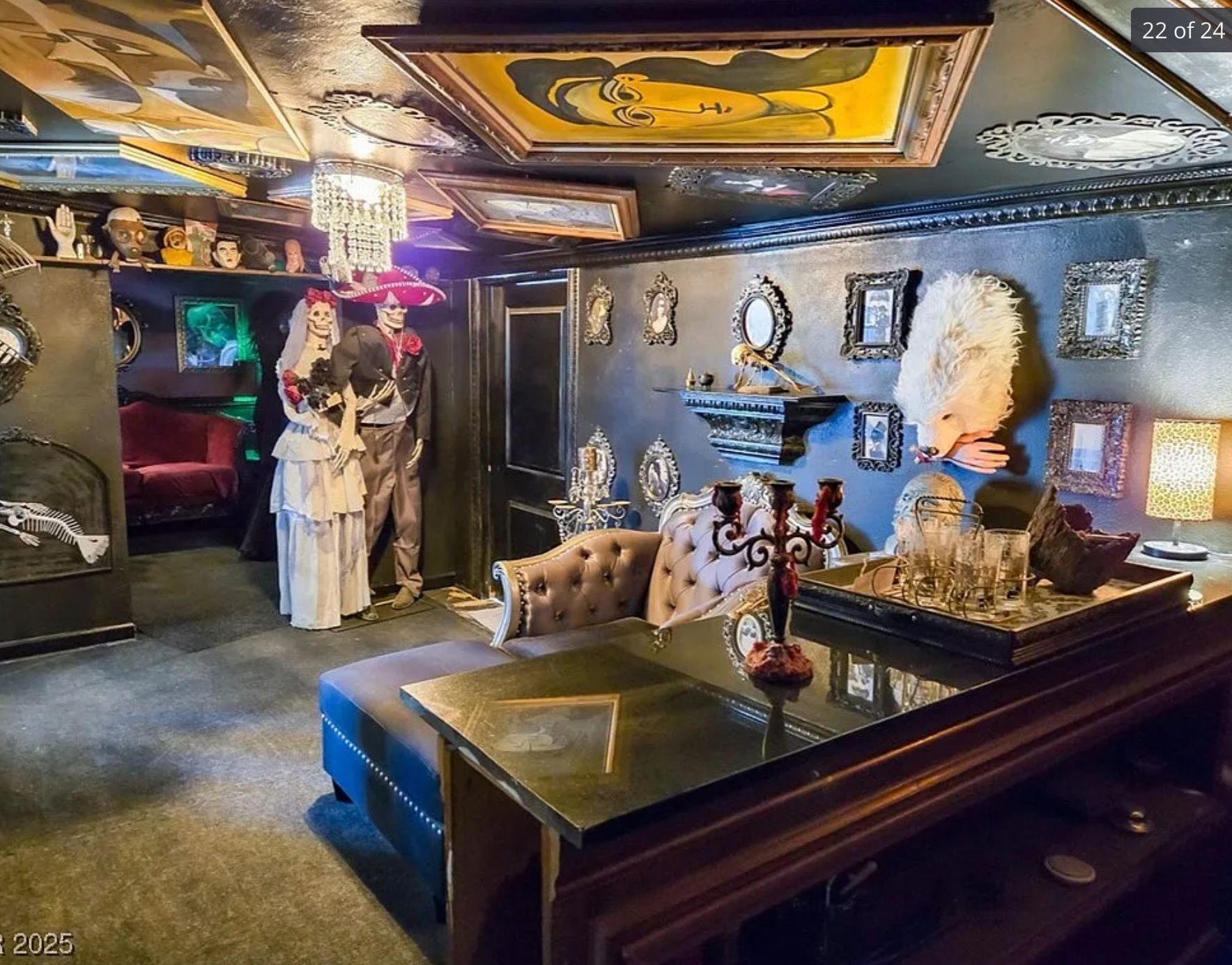 A basement room with black walls and paintings attached to the ceiling. There are a pair of skeletons dressed as a bride and groom, and a stuffed werewolf on the wall holding a fake bloody hand. 