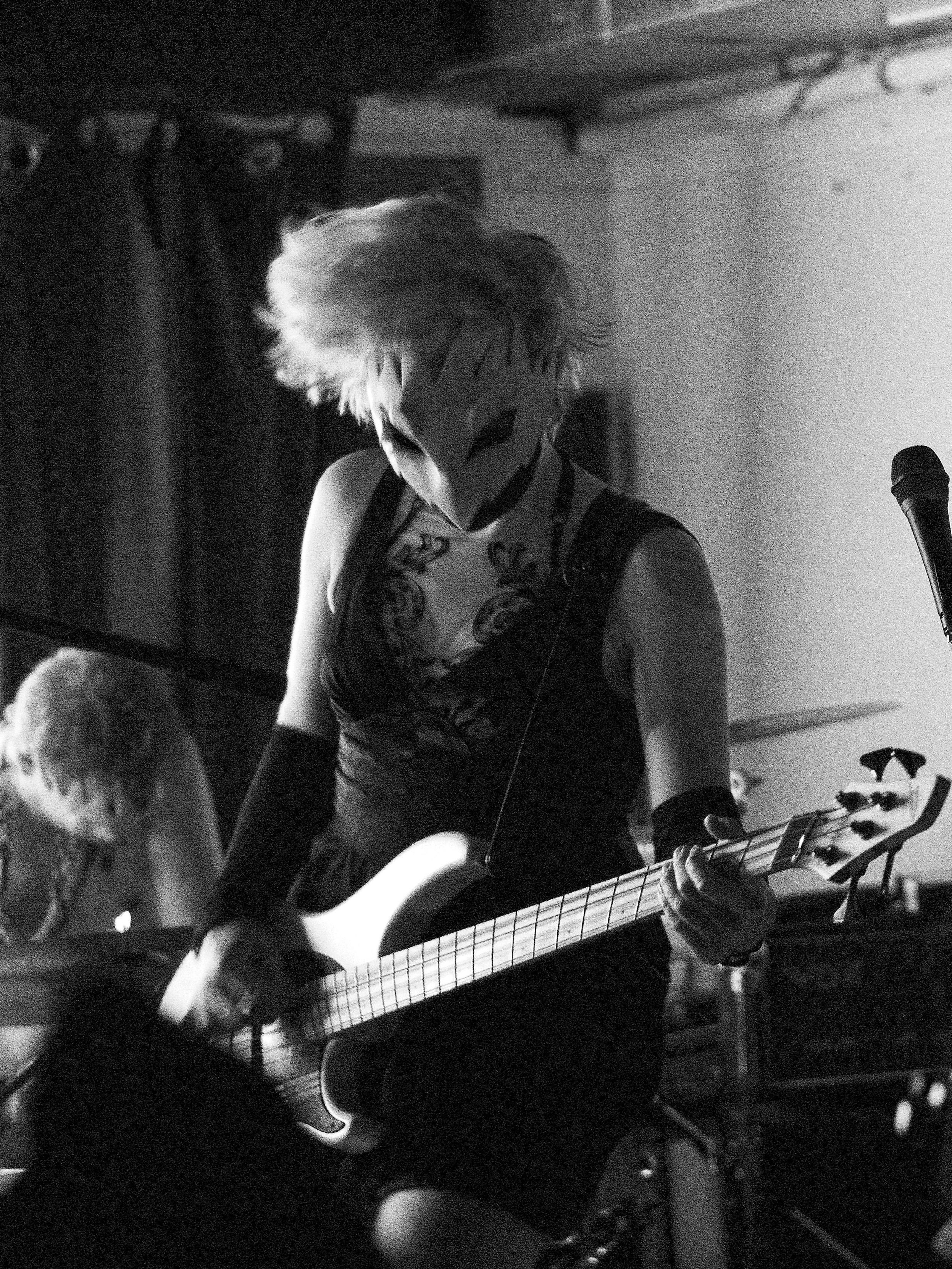A black-and-white photo of a girl with short hair on a stage playing bass guitar. She is wearing a black dress and a white birdlike mask. Photo by Orlando daSilva