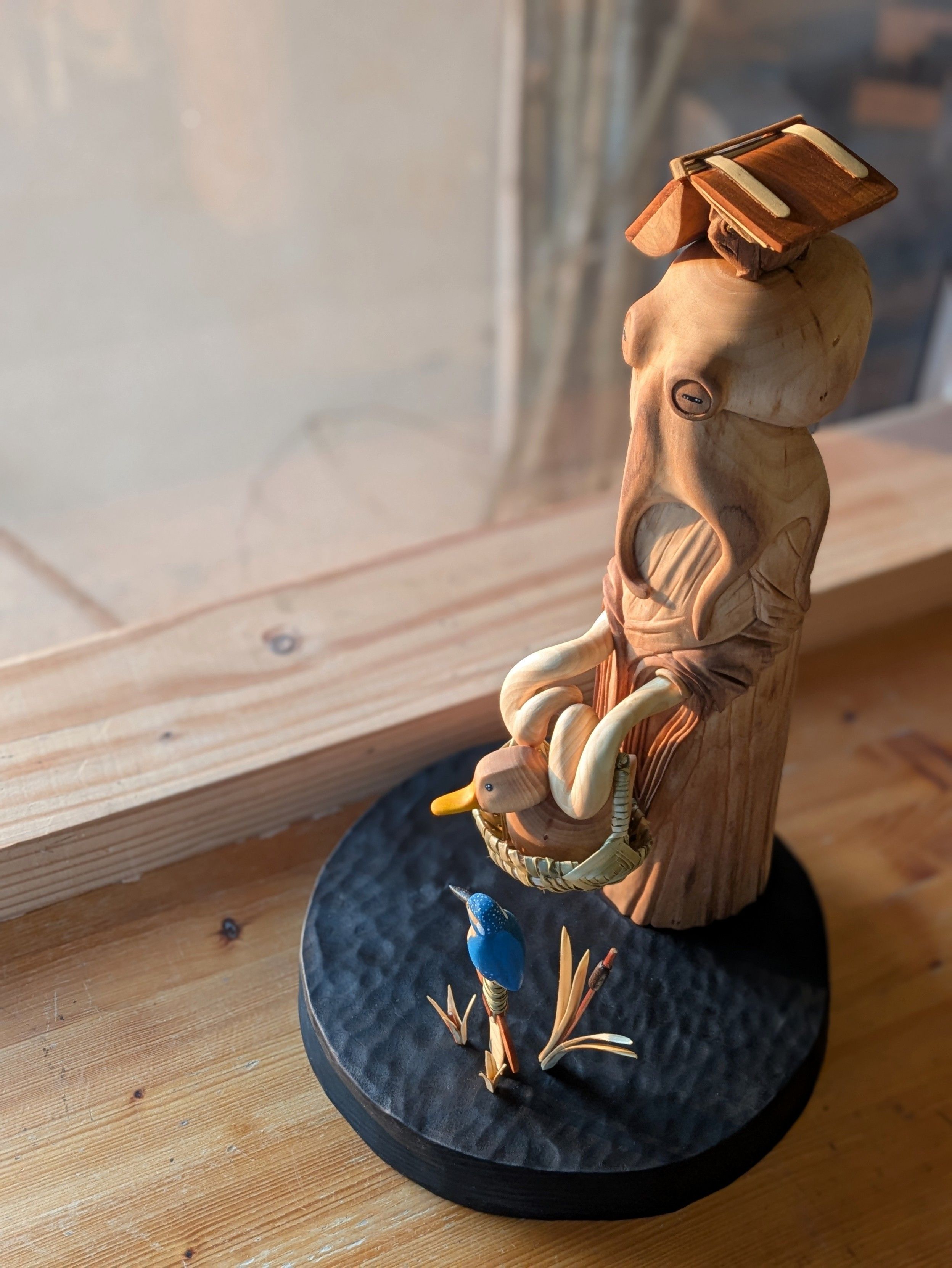 Woodcarved scene on a round base. An humanoid octopus wearing a dress is holding a basket with a duck inside of it. On the head of the octopus, a grumpy frog taking shelter of a big ancient book. Next to them all, a sword is coming out of water among bulrush. On the top of it sits a kingfisher.