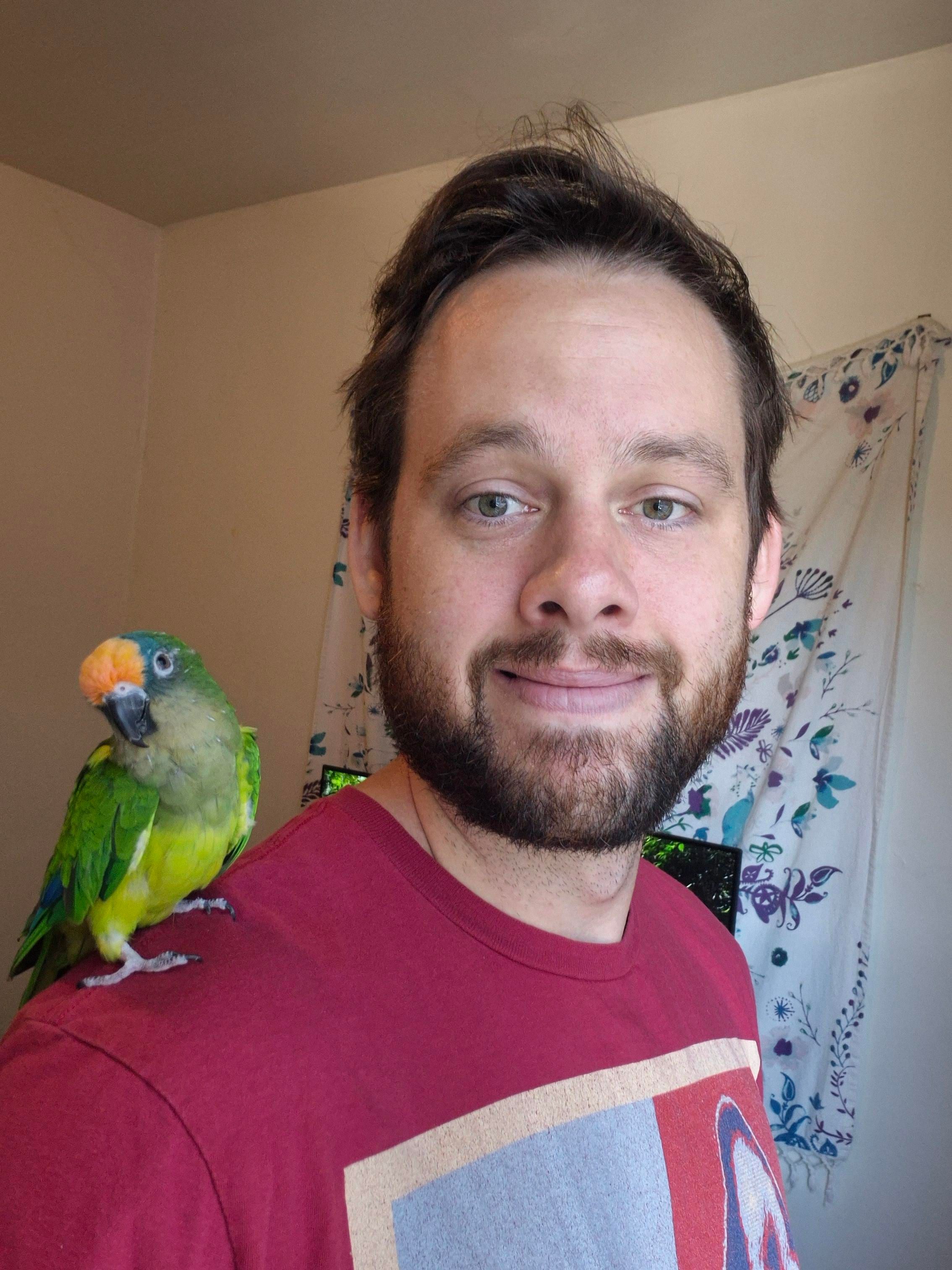 Me wearing a red shirt with a green conure on my shoulder.
