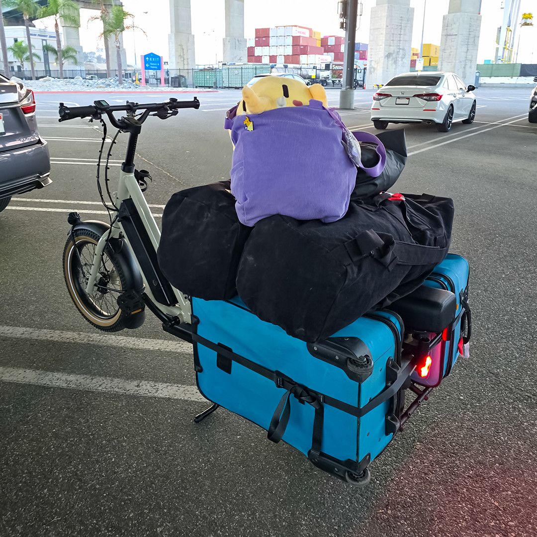 Photo of the Creature Mastodon stuffed toy sitting on a large pile of luggage strapped to the rear rack of a cargo e-bike at a harbor loading dock during one bright afternoon. The Creature is in its purple tote on top of two full black military-style duffle bags, which rest on a pair of blue suitcases ratchet-strapped together and positioned sideways on either side of the bike's running boards. The front of the cargo bike is positioned 10 o'clock to the camera, with the red rear light shining toward 4 o'clock. All around the Creature in the surrounding parking lot are parked cars, and in the distance, palm trees and shipping containers are visible.