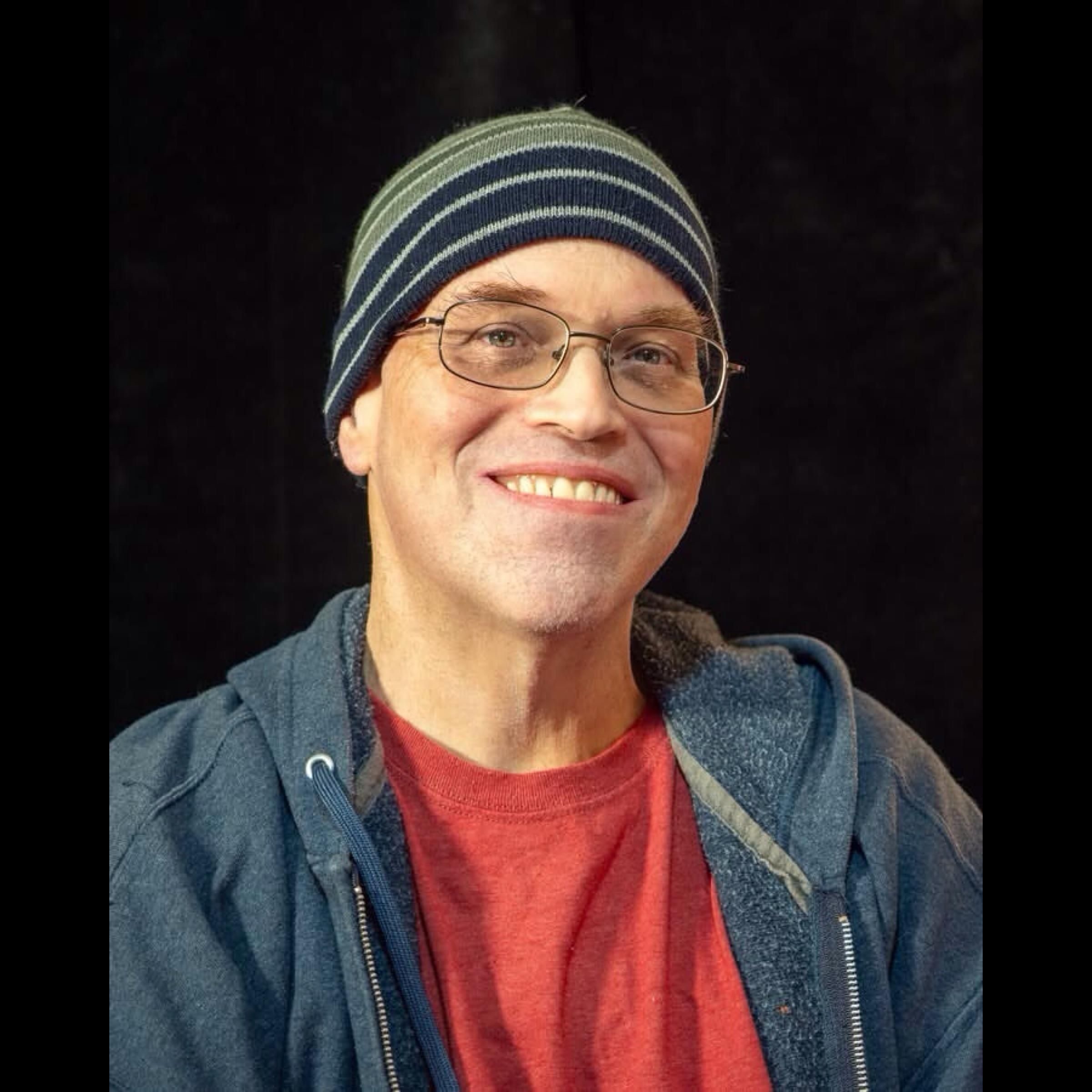 Photo of a man wearing a plain red t-shirt and an unzipped blue hoodie. He has eyeglasses and is wearing a blue striped beanie cap.
