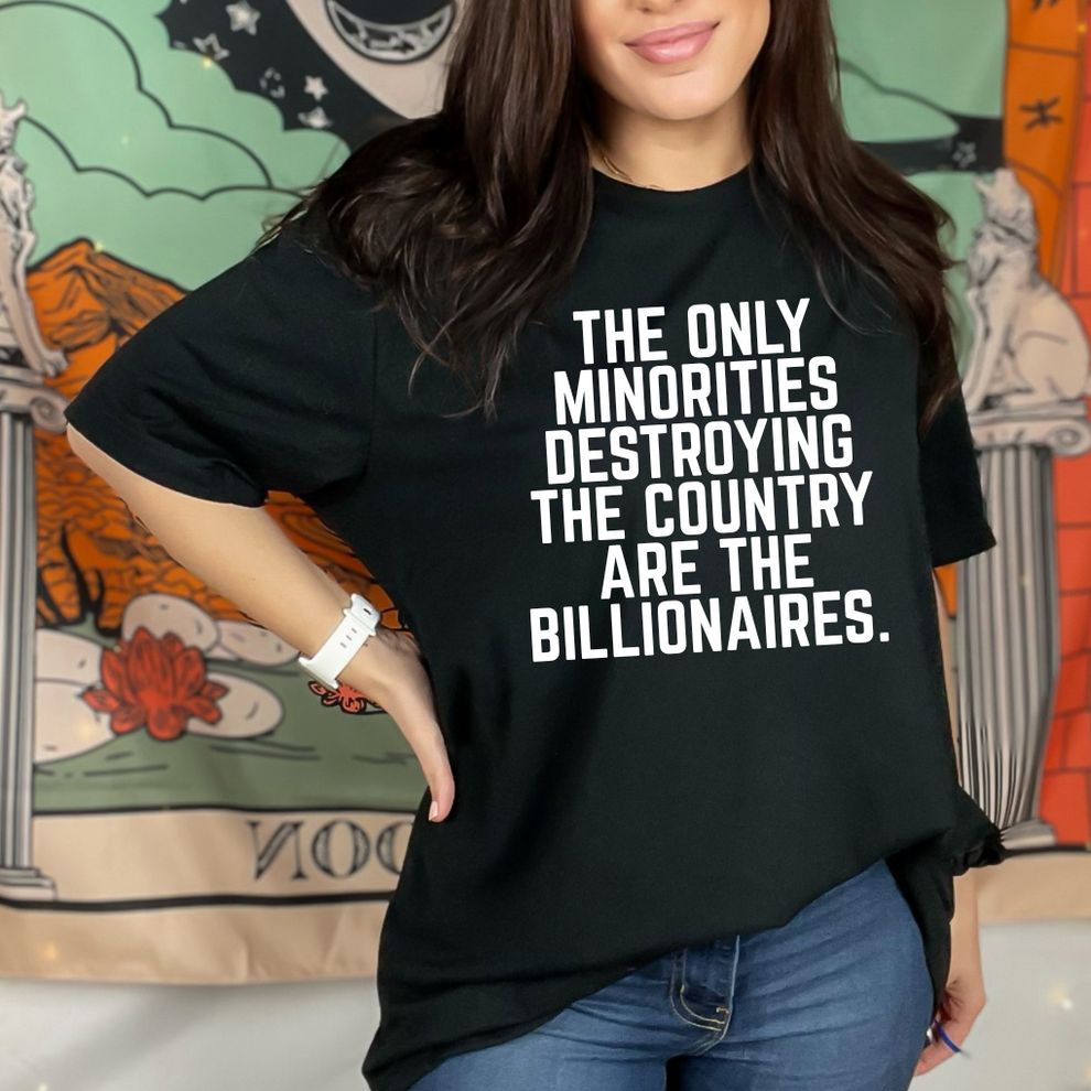A woman wears a black t-shirt with white, bold, all-caps text that reads: "THE ONLY MINORITIES DESTROYING THE COUNTRY ARE THE BILLIONAIRES." She stands with one hand on her hip against a background featuring a colorful, illustrative tapestry.
