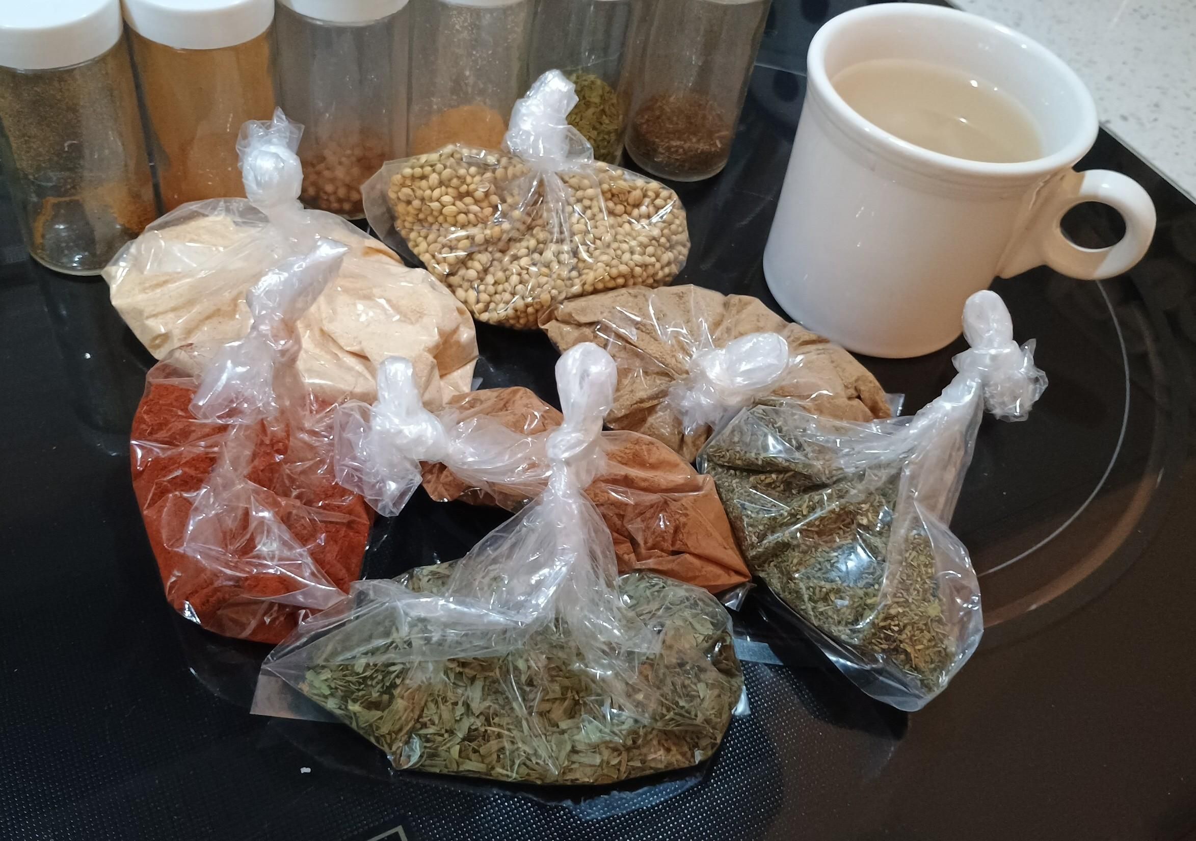 A bunch of transparent bags of the aforementioned spices, and a white mug beside them. A row of small jars is in the background and waiting to be filled.