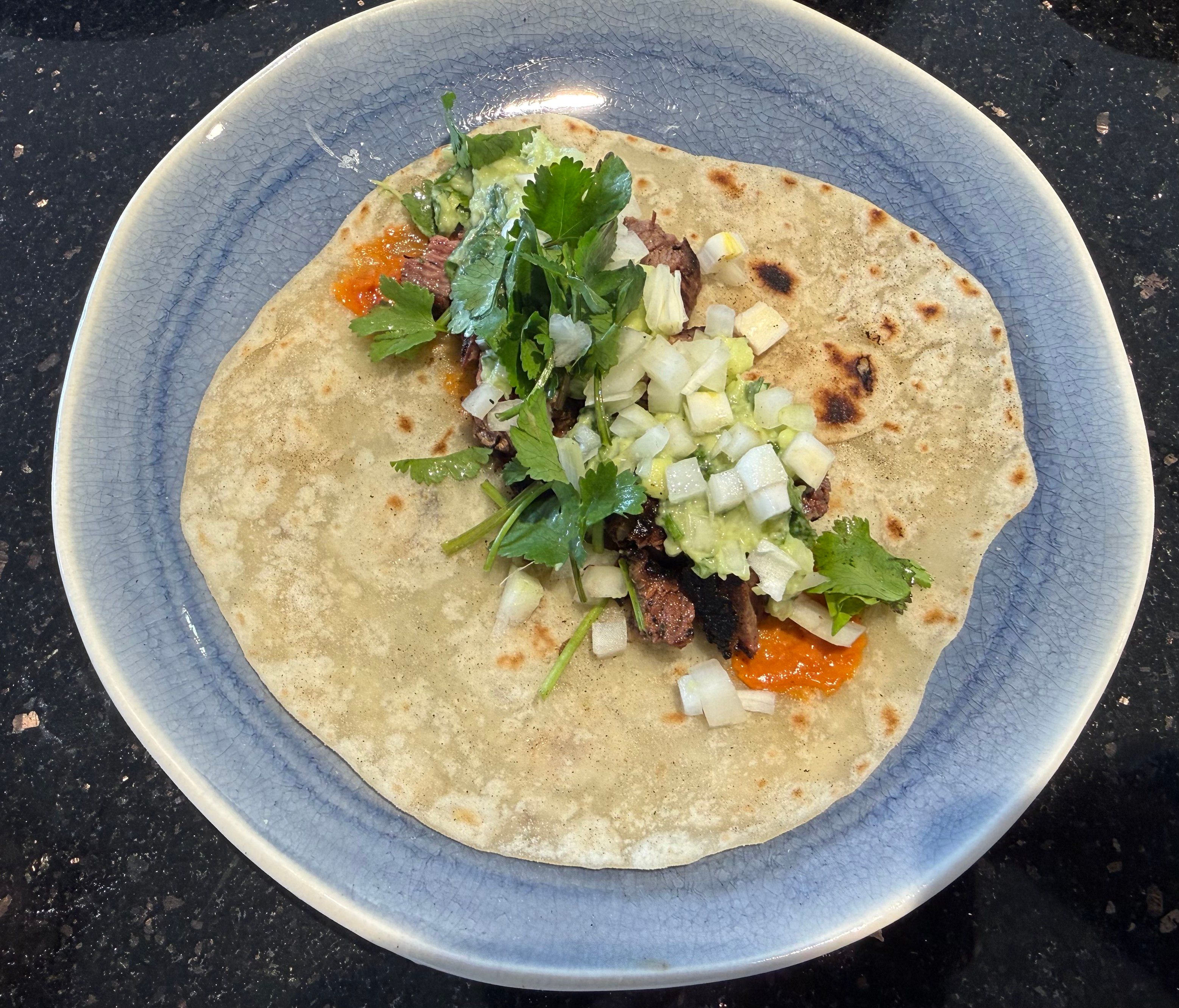 Flour tortilla with meat, onions, cilantro, guacamole and underneath a red/orange chili arbol salsa is peeking through. 