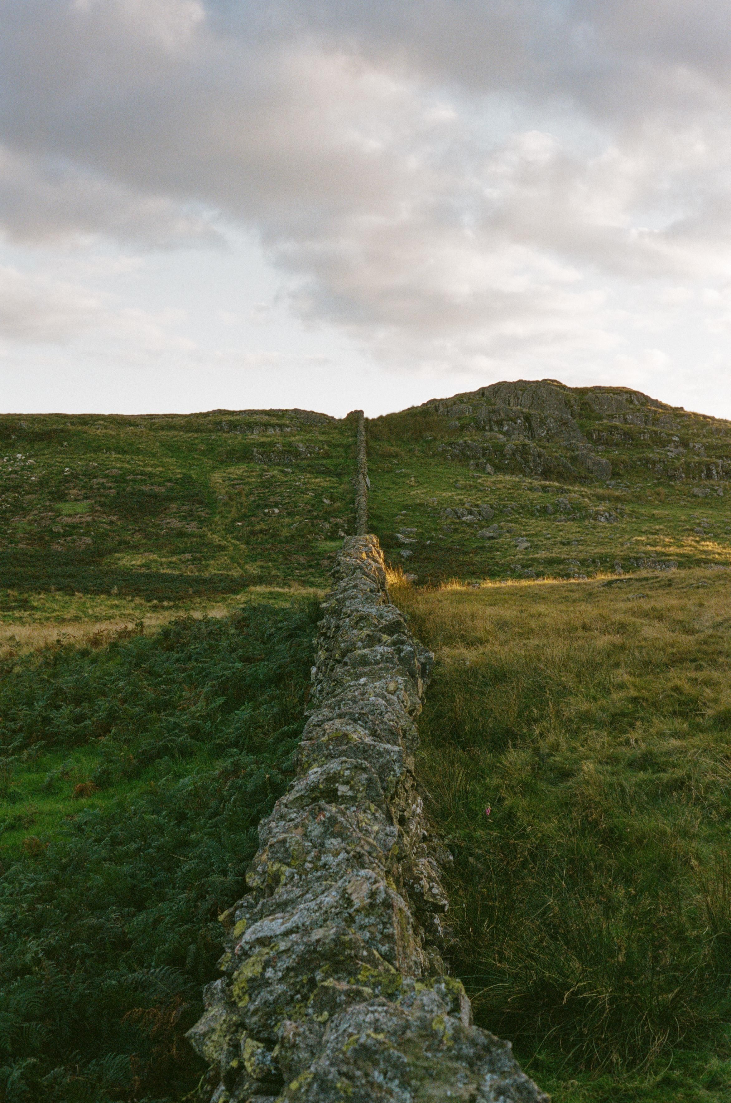 A dry-stone wall extending into the distance, splitting the green landscape into two equal parts. A ray of golden sunlight descends on the middle part.