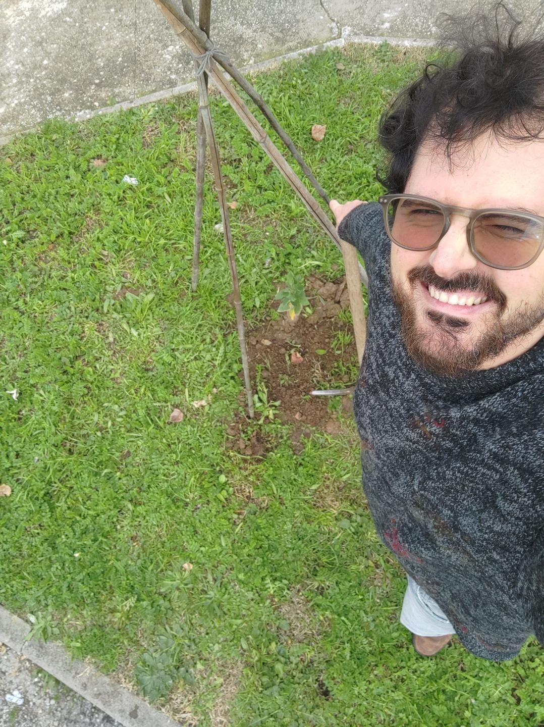 Gabriele Primavera, in un maglione marrone, appoggiato a un piccone fa un selfie con un nuovo melo piantato in un'aiuola, posto sotto una struttura di cannicci