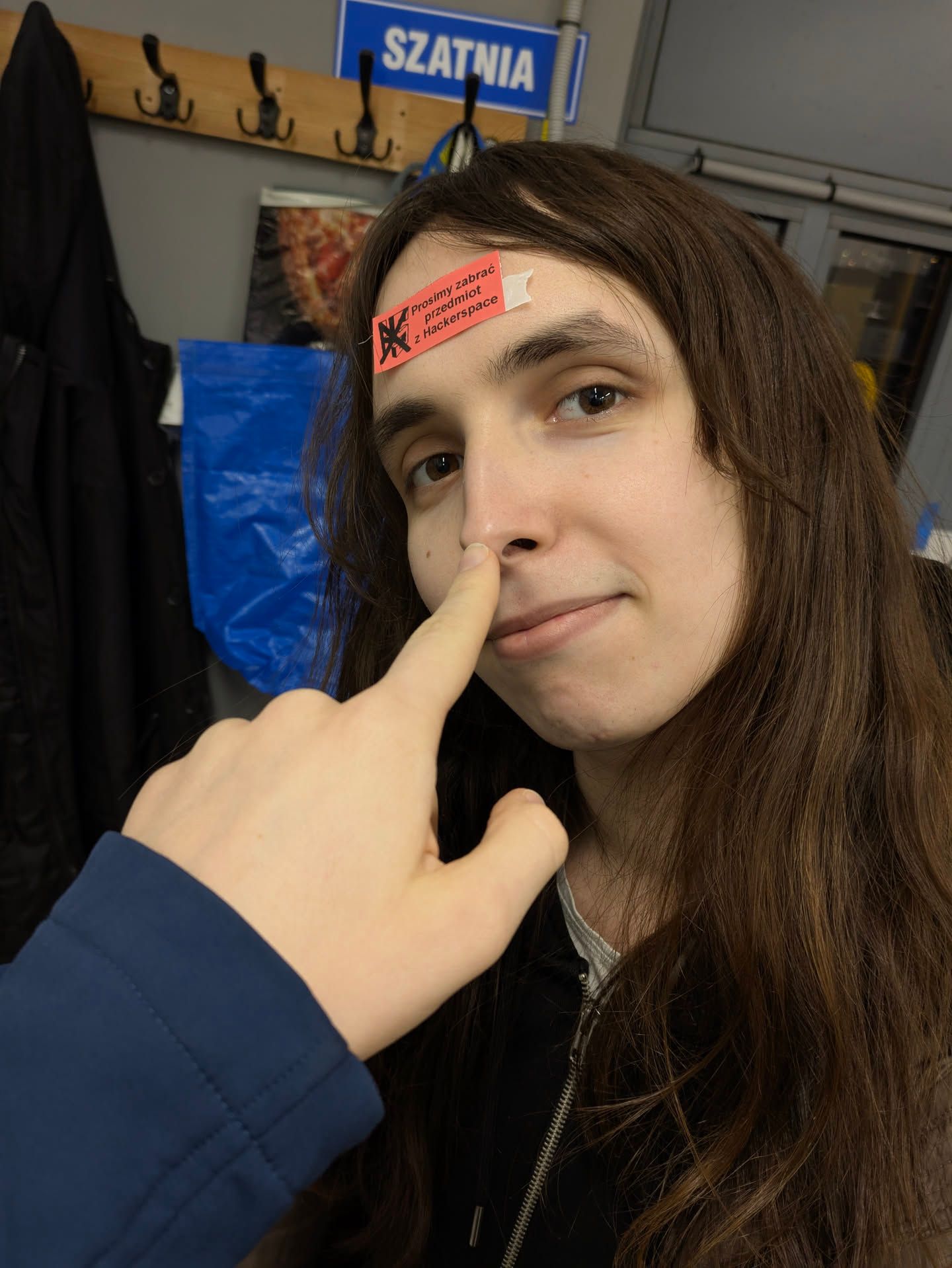 nicole, looking slightly awkwardly while being booped by fęść, with the "prosimy zabrać przedmiot z Hackerspace" (please remove [this] item from the Hackerspace) sticker on her forehead