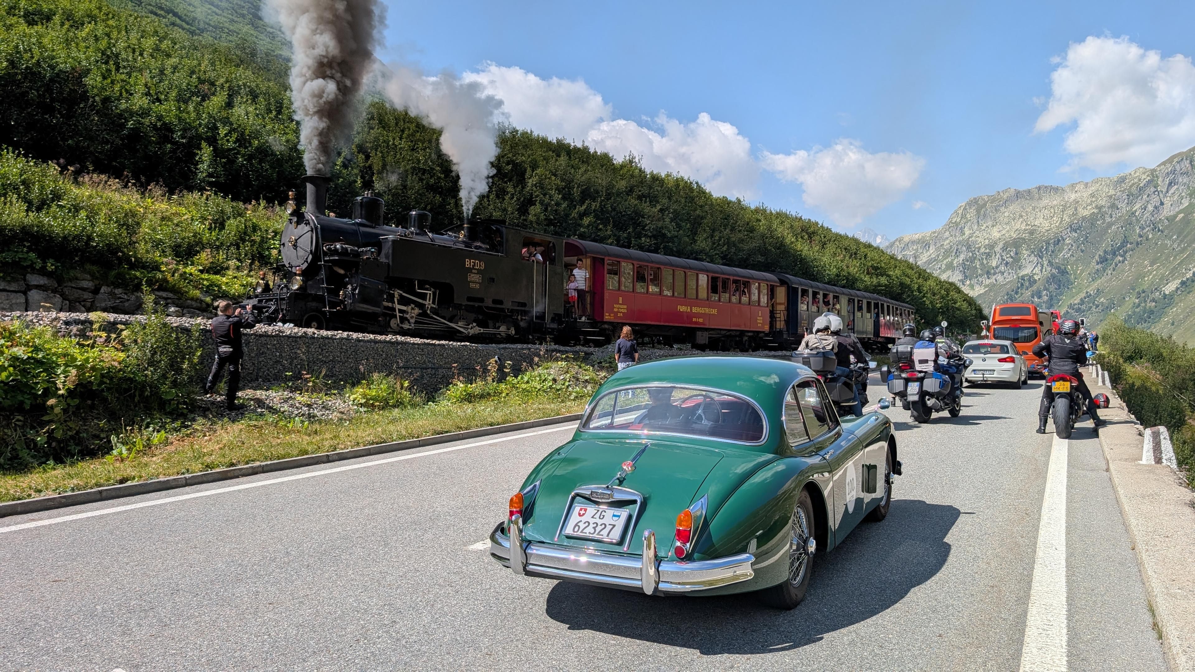 Ein Jaguar XK150 auf einer Passstrasse in den Bergen, von hinten fotografiert. Er steht gemeinsam mit anderen Fahrzeugen vor einem Bahnübergang, während ein alter Dampfzug vorbei fährt. Aus dem vorderen Schornstein steigt eine gewaltige dunkelgraue Rauchwolke. Weiter hinten an der Lok entweicht eine weisse Dampfwolke.