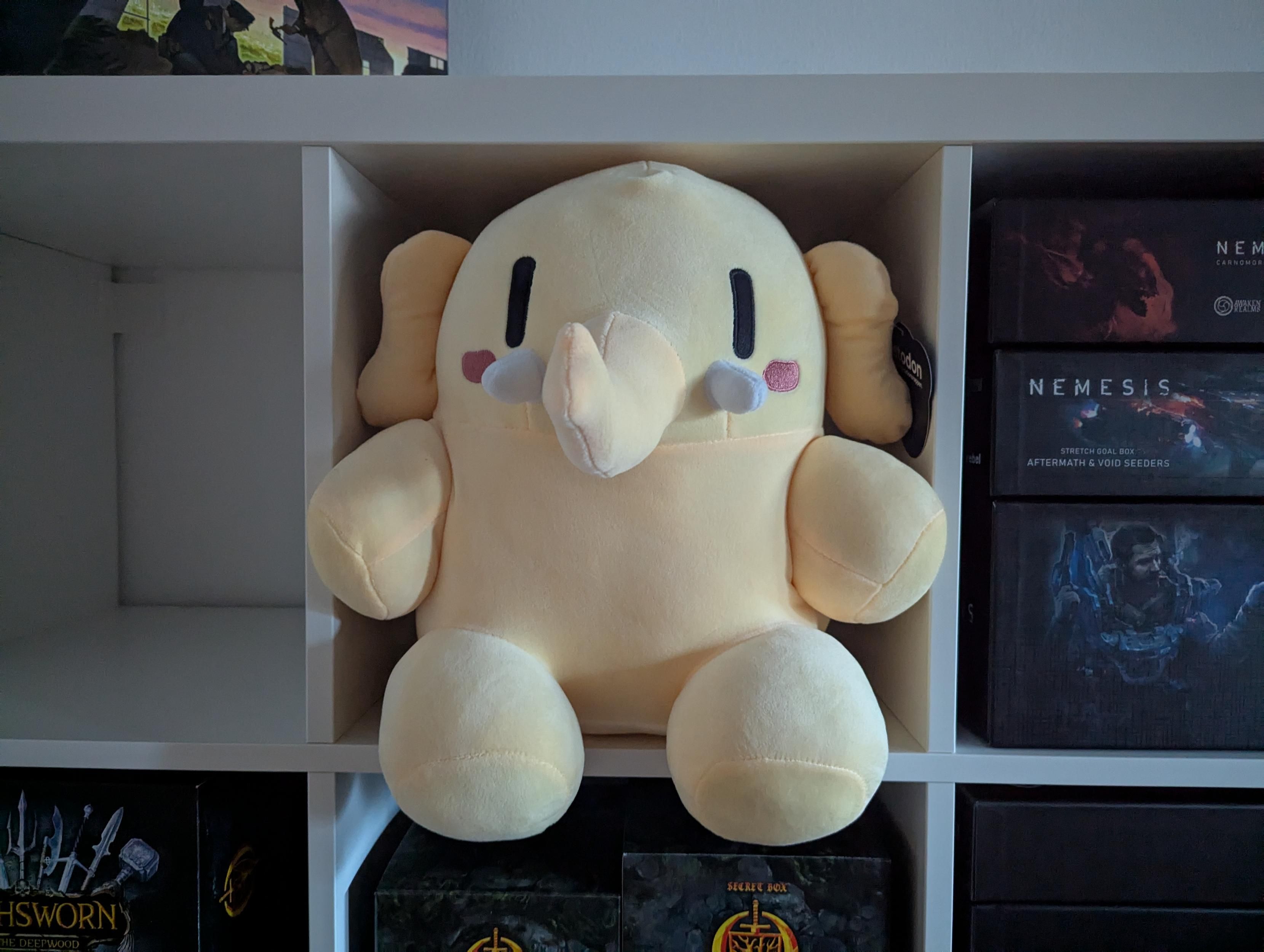 John Mastodon (my Mastodon mascot plushie) sitting comfortably and snugly in a Kallax compartment, boardgames in the compartments around it