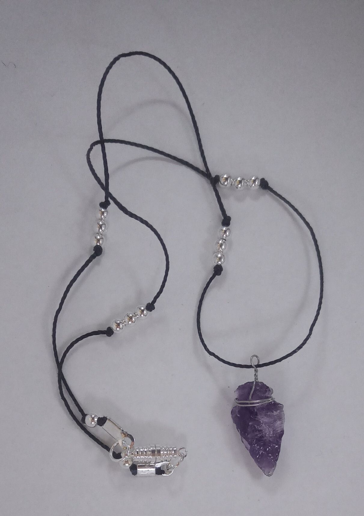 An amethyst arrowhead pendant, on a white background; the arrowhead has been knapped, so it has lots of rough, chipped-looking edges. It's strung on a black cord, with groups of 3 round silver beads at short intervals, and a silver-finished oblong magnetic clasp. The arrowhead has some silver wire wrapped around the widest point, forming a loop above it, so that it can hang from the cord.