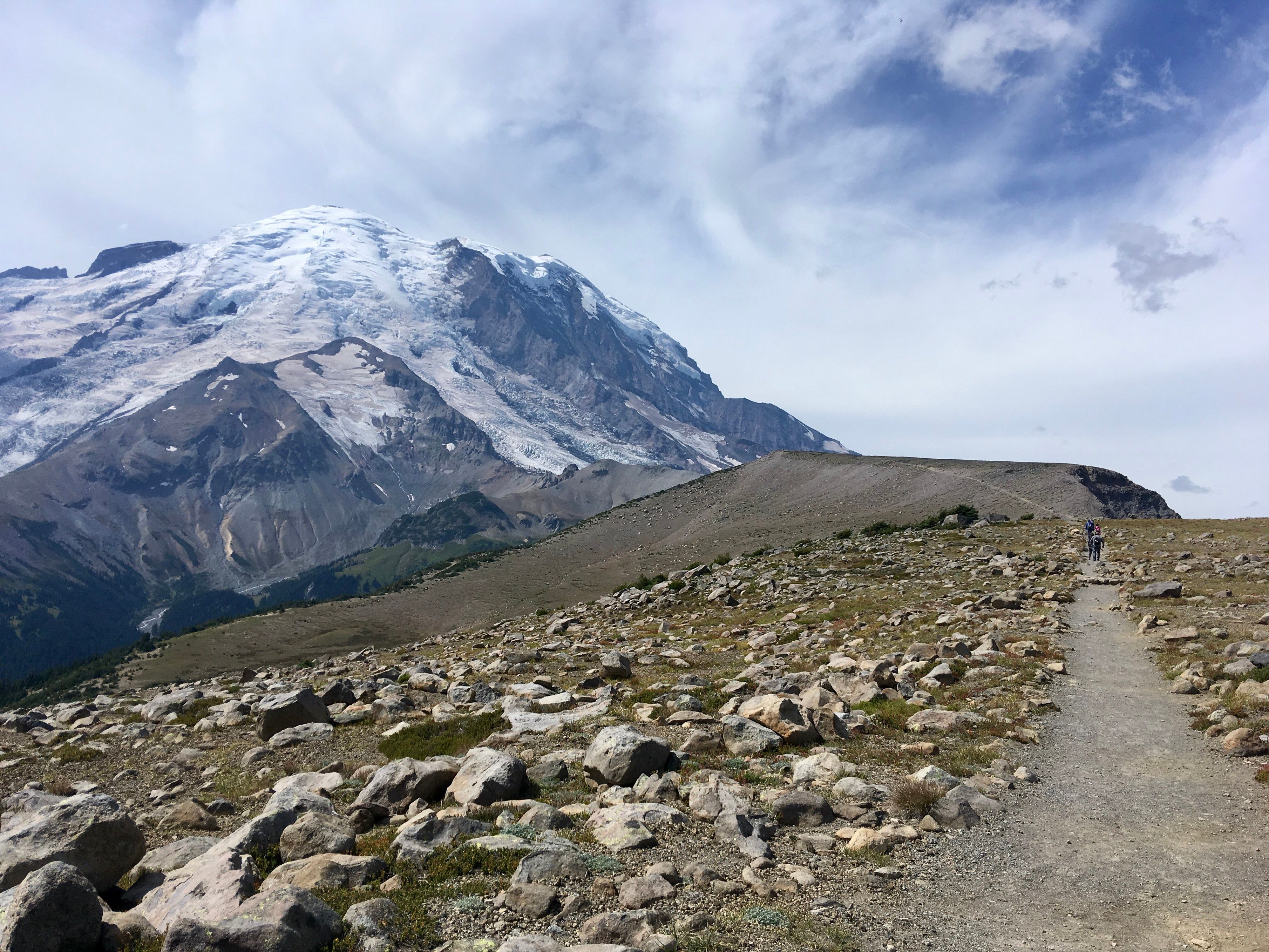 A glacier domed volcanic mountain with black underparts and wisps of copper divots loomos above a rock strewn plateau with a wide, flat path heading towards a tilted flat peak. 