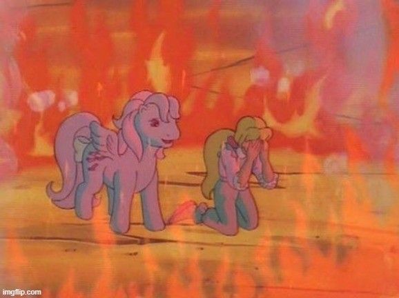 A frame of a My Little Pony cartoon. A human girl is surrounded by flames, on her knees crying, with a cheerful-looking pony approaches her.