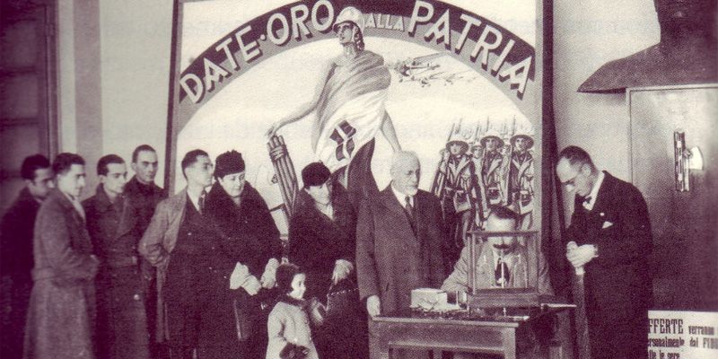 Foto dell'epoca che ritrae alcune persone in fila per donare l apropria fede nuziale. Più avanti un banchino con una bilancia e una persona che riceve le fedi. Sullo sfondo un'immagine dell'Italia personificata, vestita della bandiera Italiana, che sorregge un fascio littorio; sulle sfondo truppe armate di moschetto. Sull'immagine campeggia la scritta "Date Oro all'Italia"