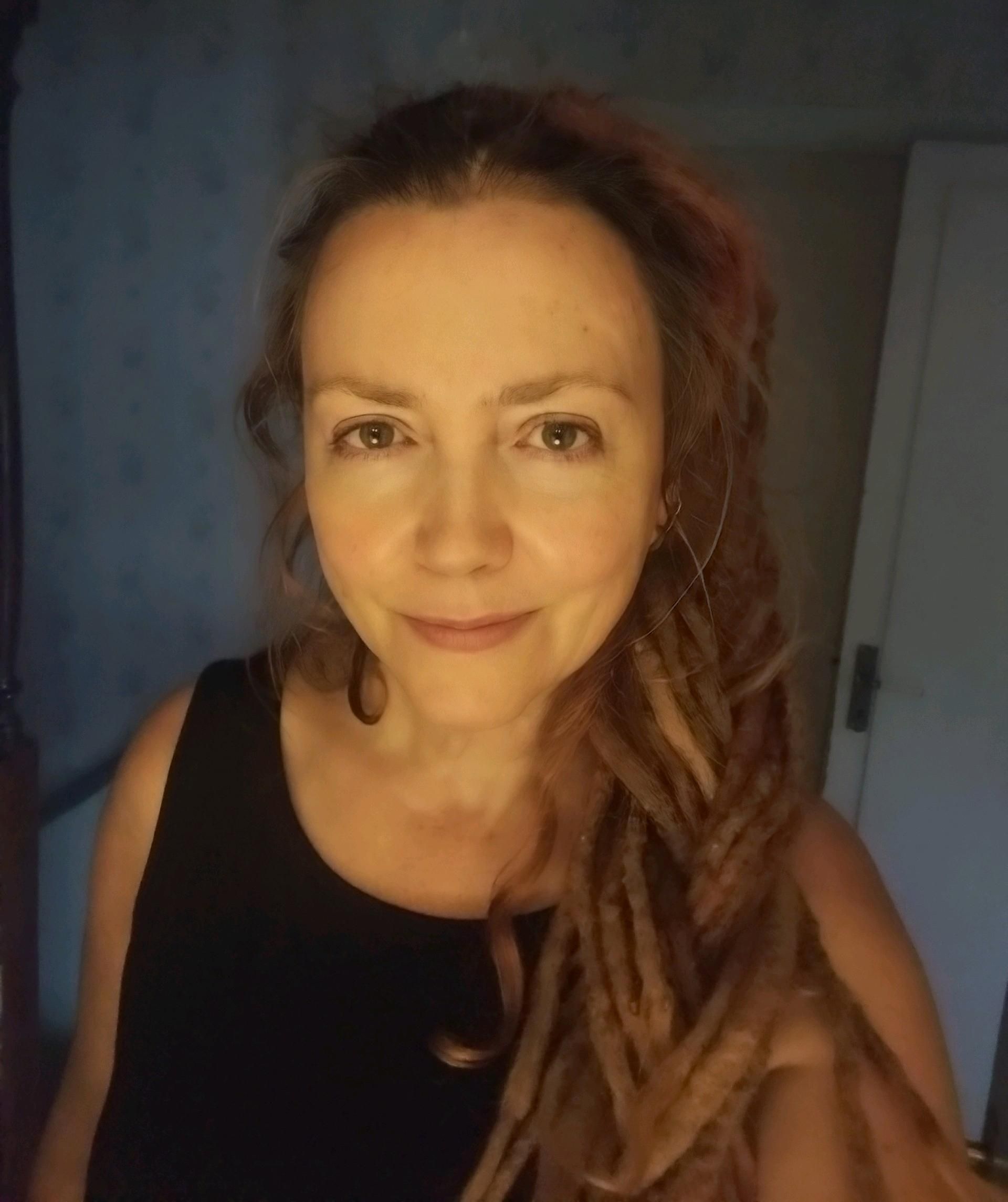 Head and shoulders shot of a white woman with brown eyes and blonde dreadlocks in a messy plait. She's looking into the camera. The light on her face is warm and golden, but it's clearly an indoor photo.