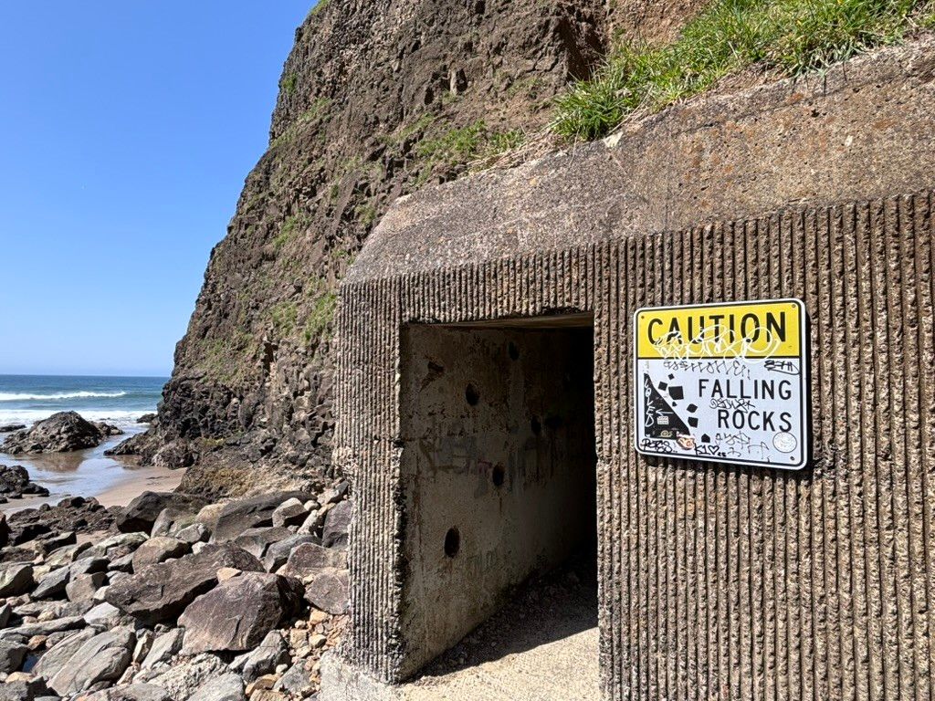 Concrete tunnel entrance embedded in a rocky cliff by the ocean, with a graffiti-marked sign warning "Caution Falling Rocks."