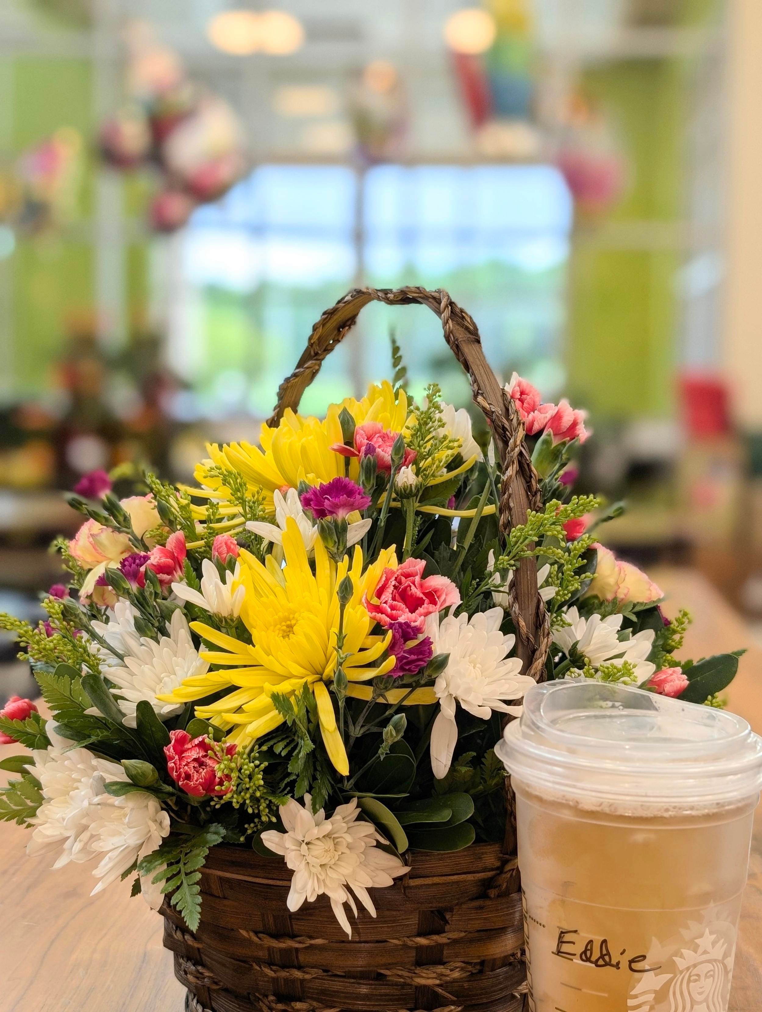 a wicker basket with a variety of flowers, including mums and carnations, sits next to a clear cup of frosty, green teaa on a long wooden table. The background is out of focus, balloons and plants in front of large double+height windows to the outside and the cloudy afternoon.