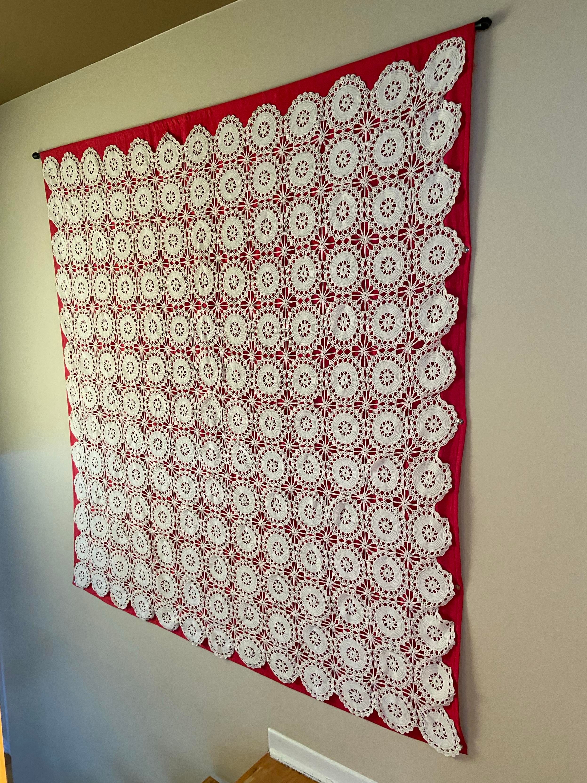 A cream colored crocheted bedspread, attached to a red backing fabric, hanging on a beige wall. We see it from an angle because it's over some stairs. 
